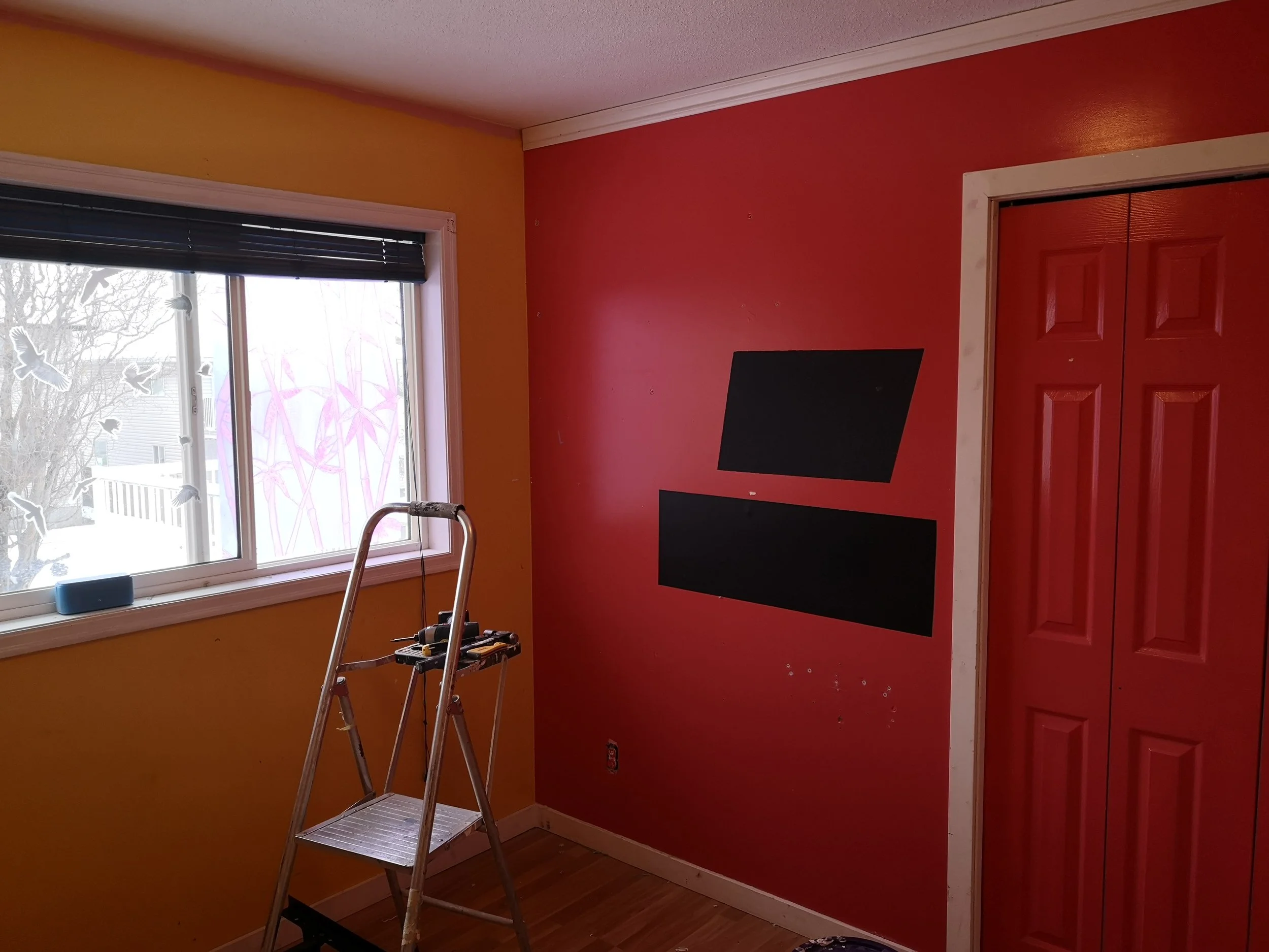 A room with yellow and red walls, a window with blinds, a step ladder, and tools, indicating ongoing painting or home improvement work.