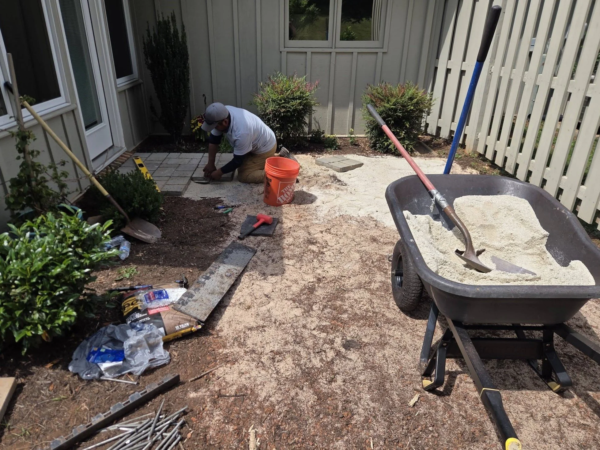 A person kneeling on the ground installing pavers in a patio area next to a house. Various tools and materials such as a wheelbarrow filled with sand or gravel, a level, a hammer, and a bucket are visible around them. The patio is being built next to a garden with bushes and a white fence.