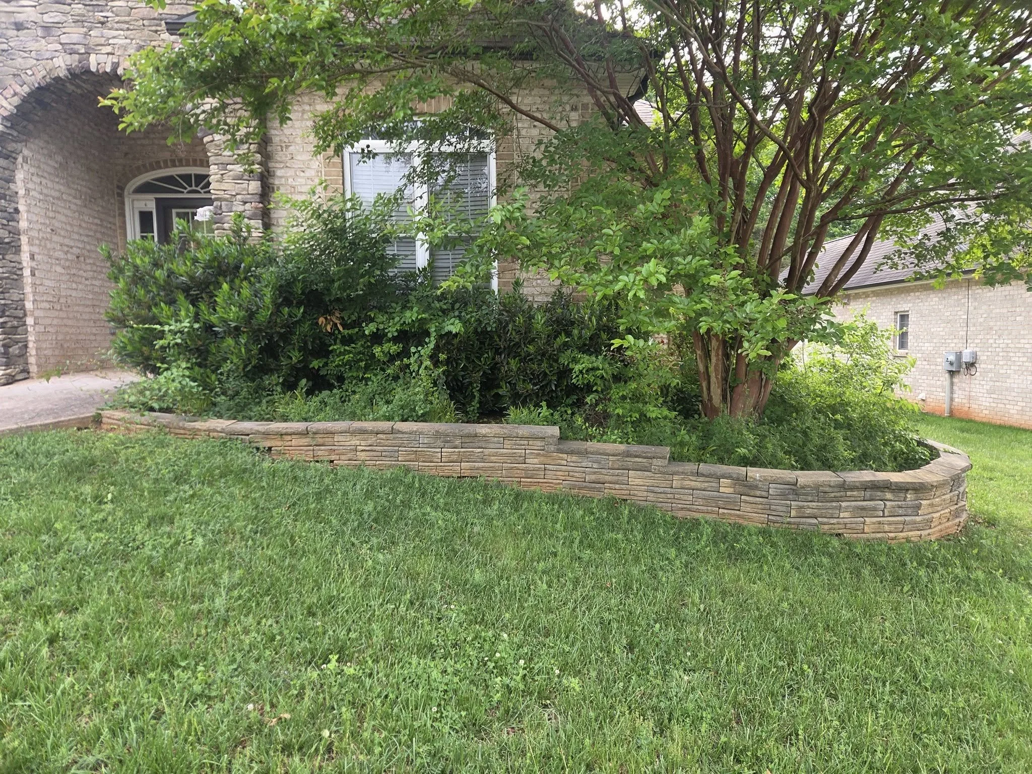 A front yard with lush green grass, bush, and a large tree in front of a brick house with a window.