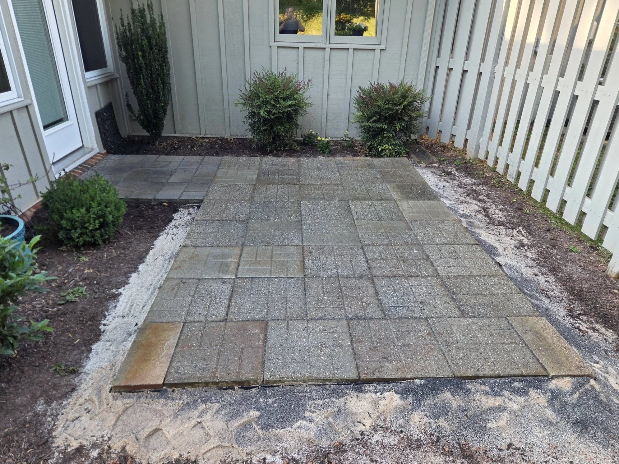 Photo of a small patio with gray pavers, surrounded by plants, and with a white fence on the right side.