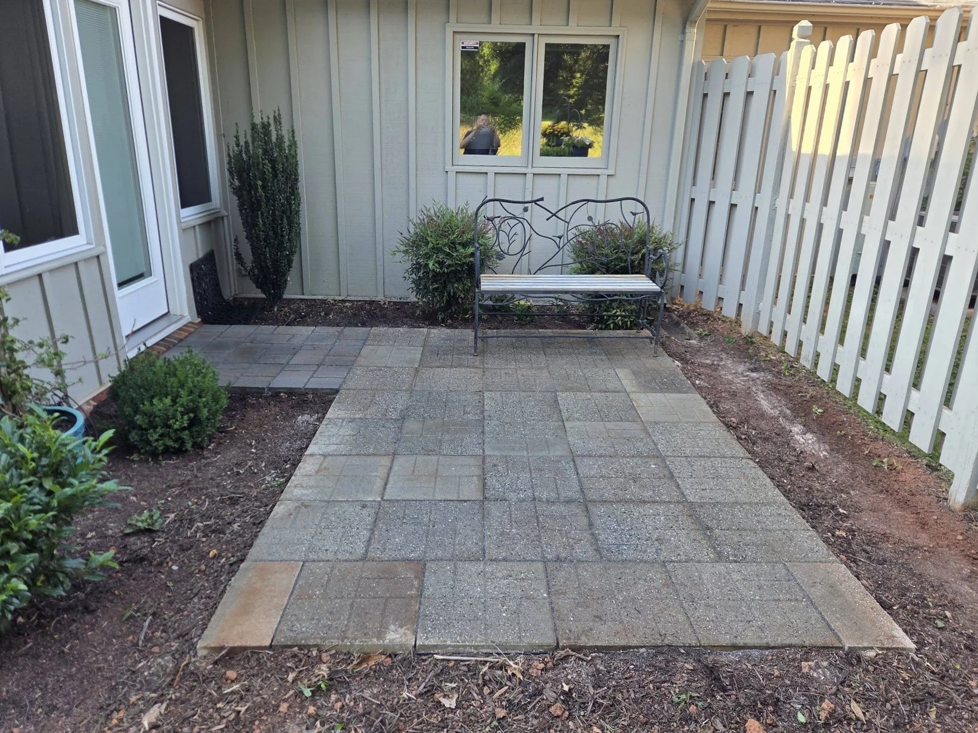 Backyard patio with a paver stone walkway, a white wooden fence on the right, a gray house wall with a window, and a decorative garden bench with plants on either side. There are shrubs and plants along the edges.