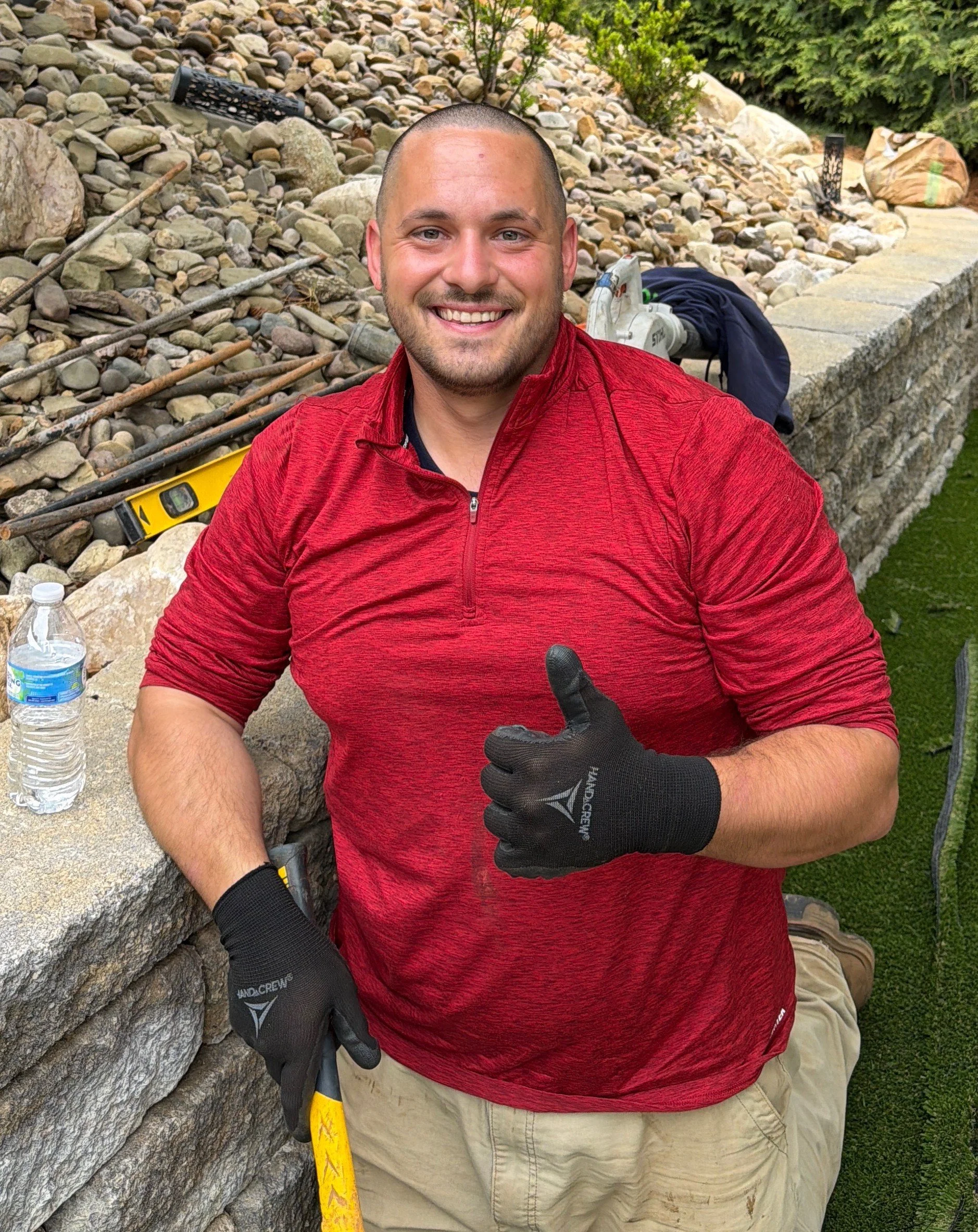 Smiling man with a buzz cut hair wearing a red athletic shirt and beige shorts, giving a thumbs up while sitting outdoors next to a stone wall with tools and water bottles nearby.