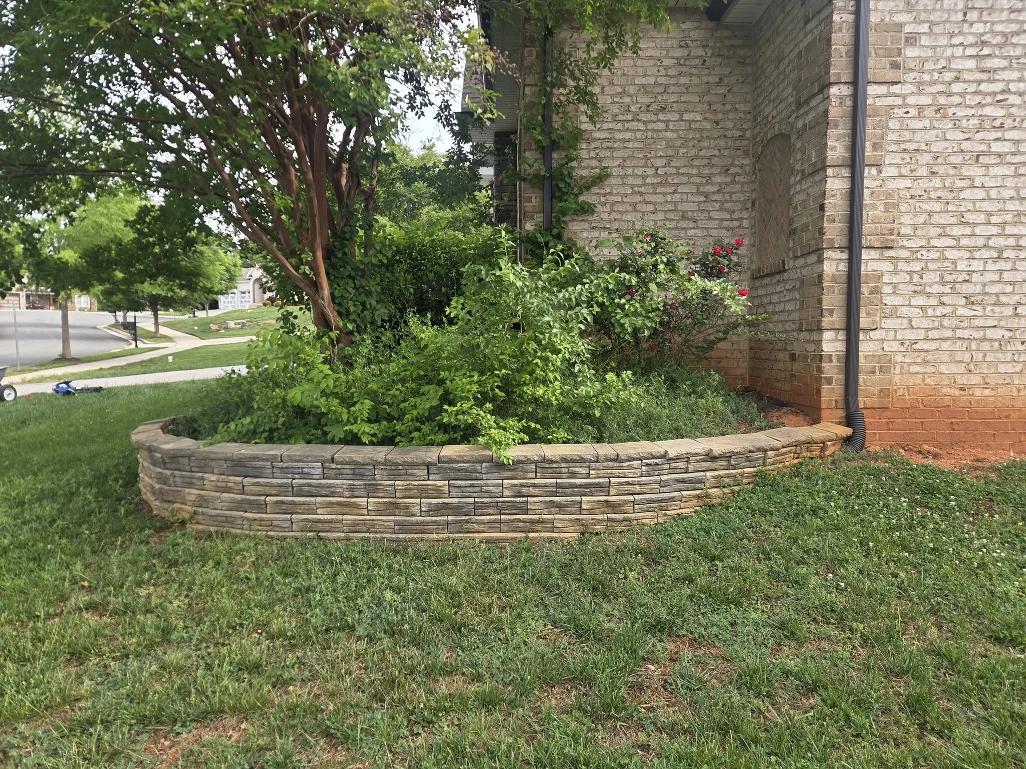 A brick house with a garden bed surrounded by a stone border, a tree, and a lawn in a suburban neighborhood.