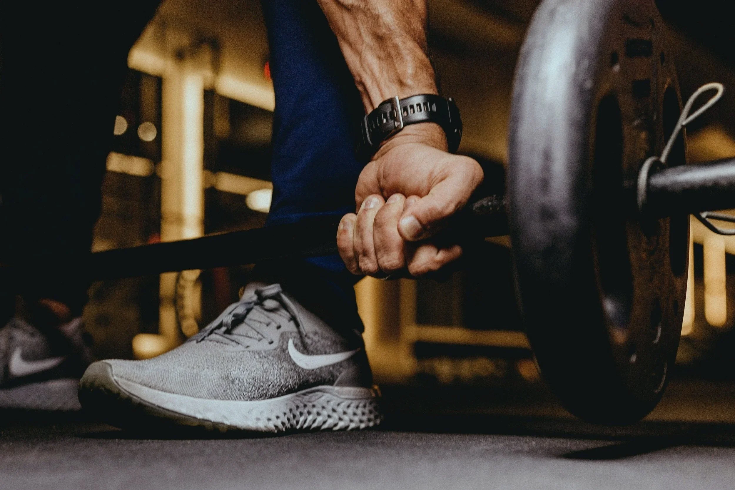 Close-up of an elderly man's hand gripping a barbell during weightlifting, wearing a black wristwatch and gray athletic shoes with white Nike swoosh, in a gym setting.