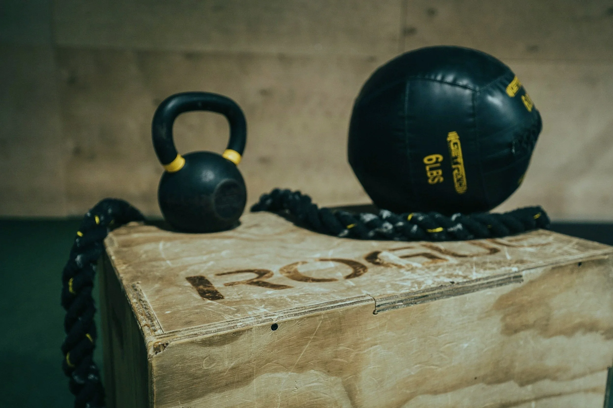 A wooden workout box with the word 'PRO' written on it, a black kettlebell with yellow accents, a black and yellow medicine ball, and a black battle rope with yellow accents, placed on a flat surface.