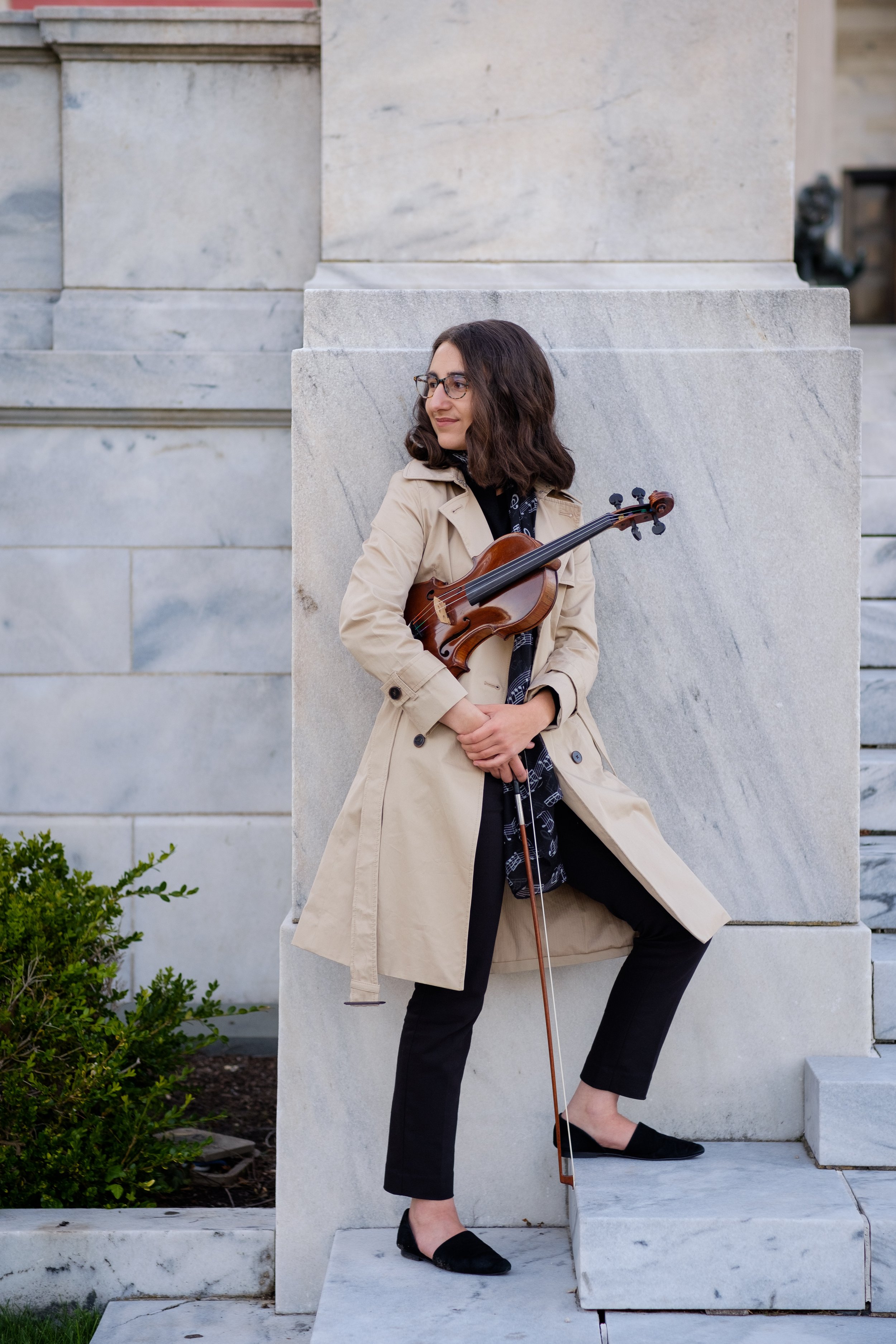 A woman with dark wavy hair, glasses, and a beige trench coat holding a violin and bow, standing outdoors against a large stone structure, with green plants and steps nearby.