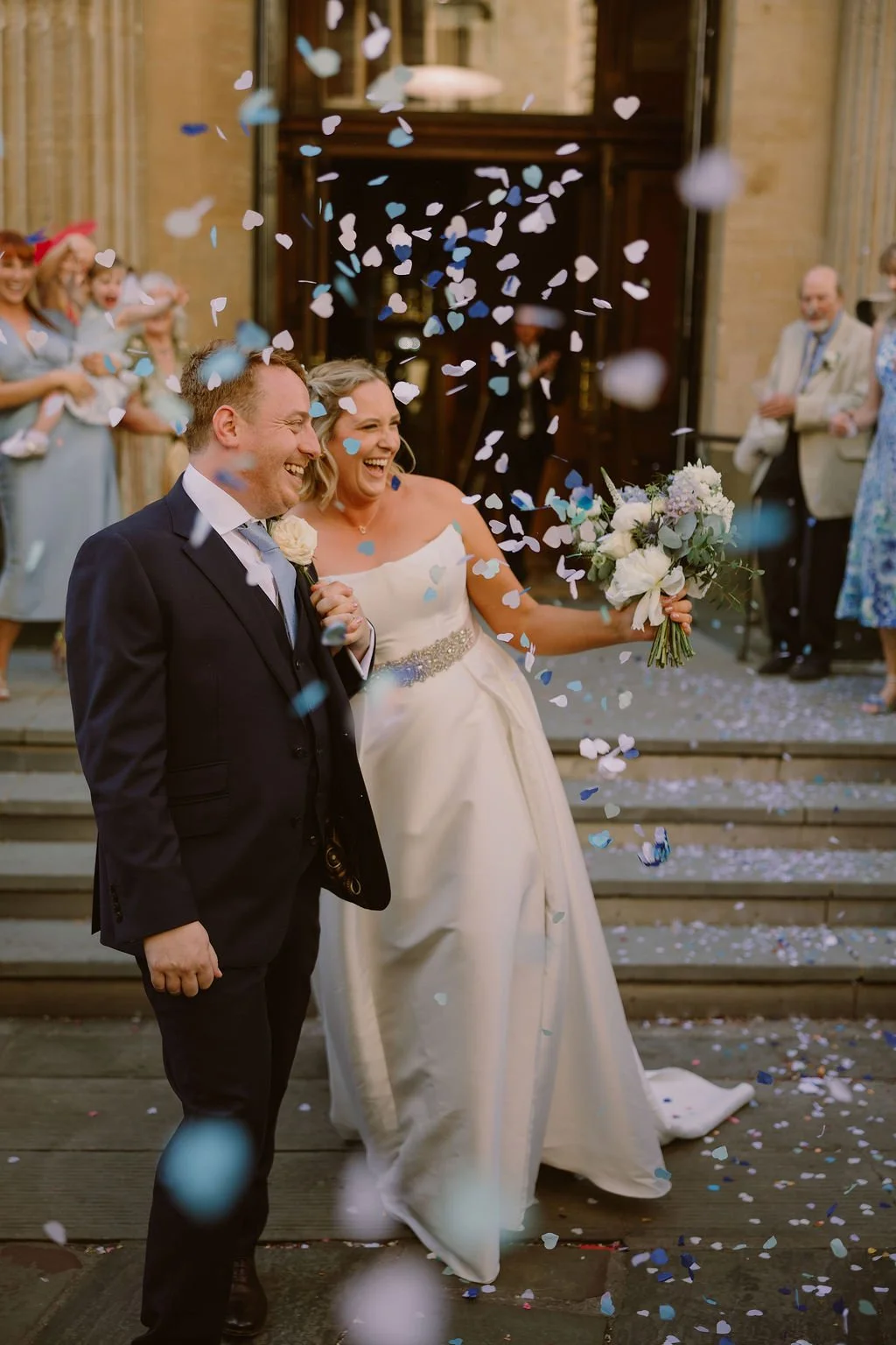 A newly married couple exiting a church, smiling and celebrating with confetti being thrown in the air. The bride is holding a bouquet of white flowers, and the groom is dressed in a dark suit with a white shirt and light blue tie. Guests are seen on the steps clapping and smiling in the background.