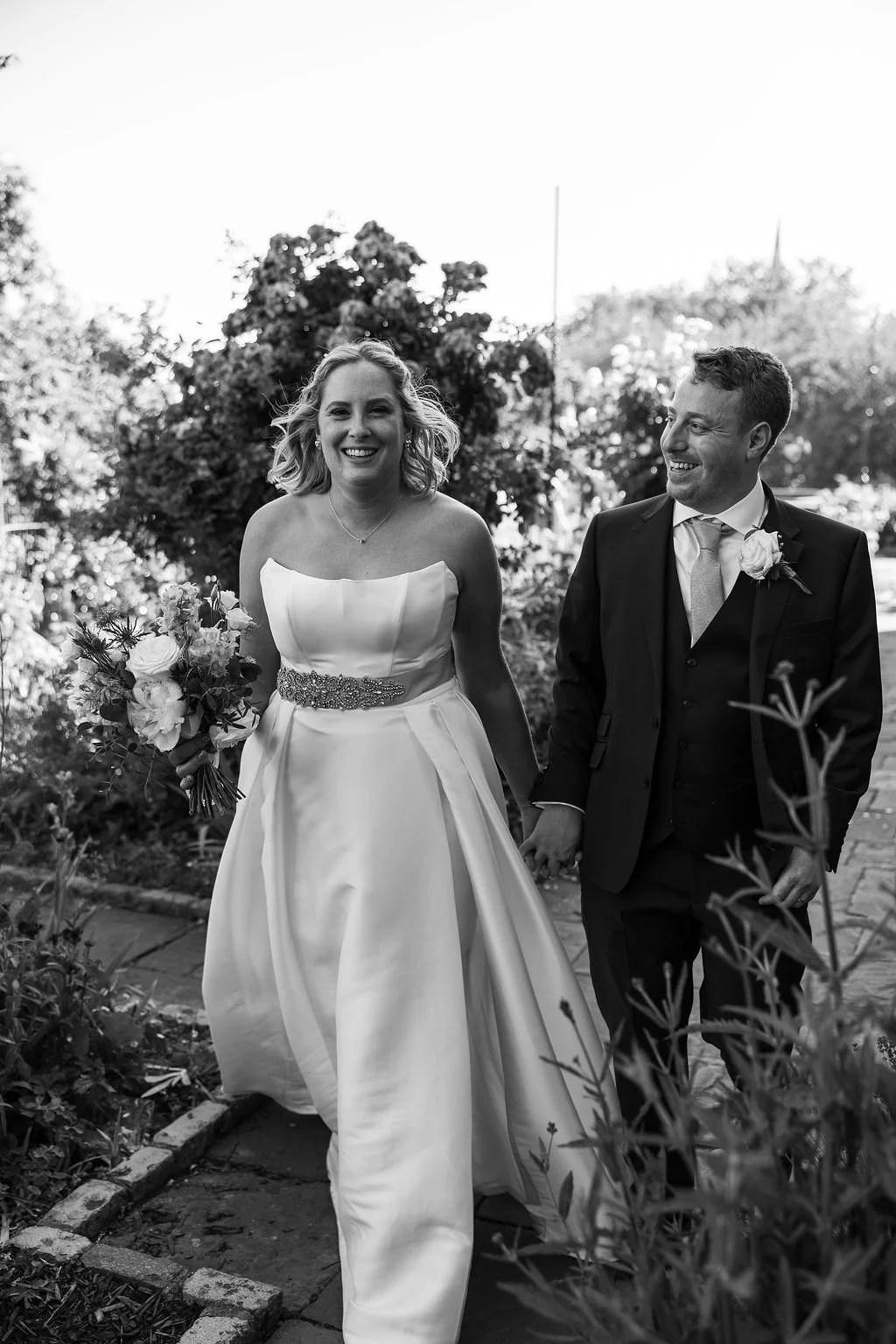 A bride and groom smiling and holding hands outdoors on a wedding day.