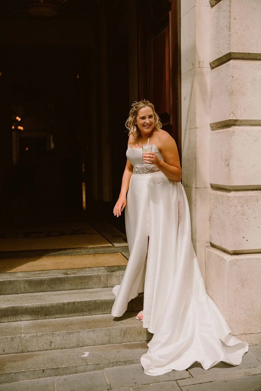 A smiling bride in a white wedding gown standing on steps outside a building at night, holding a drink with a straw.
