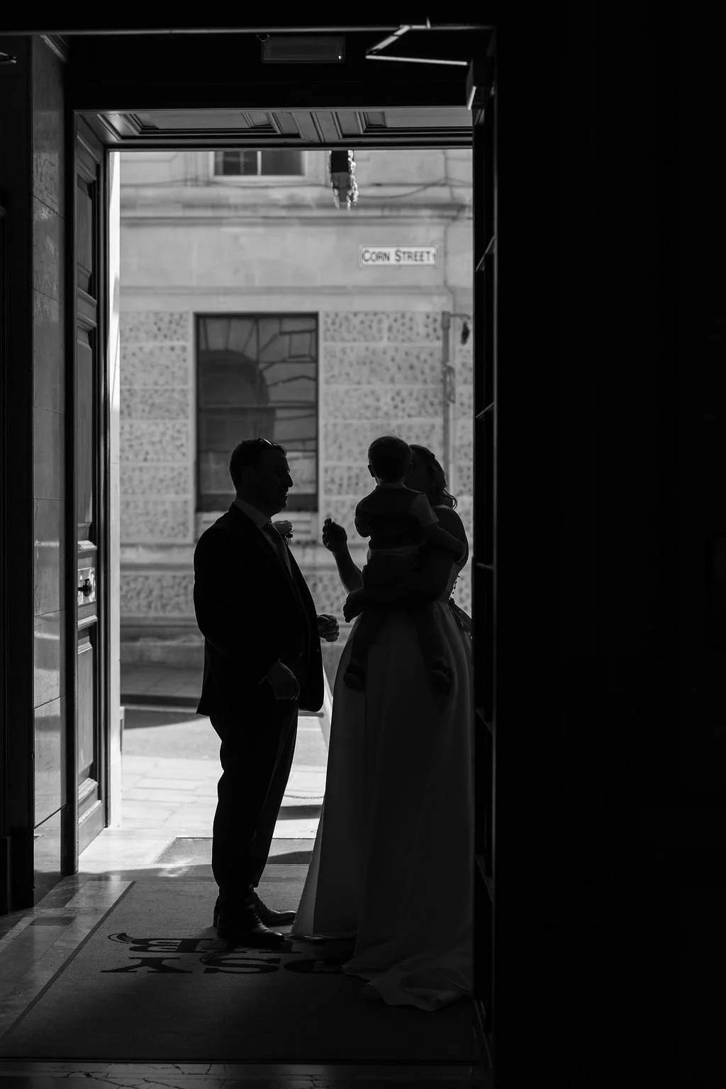 Silhouettes of a man, woman, and child standing in a doorway, with a street sign reading 'CORN STREET' visible outside in the background.