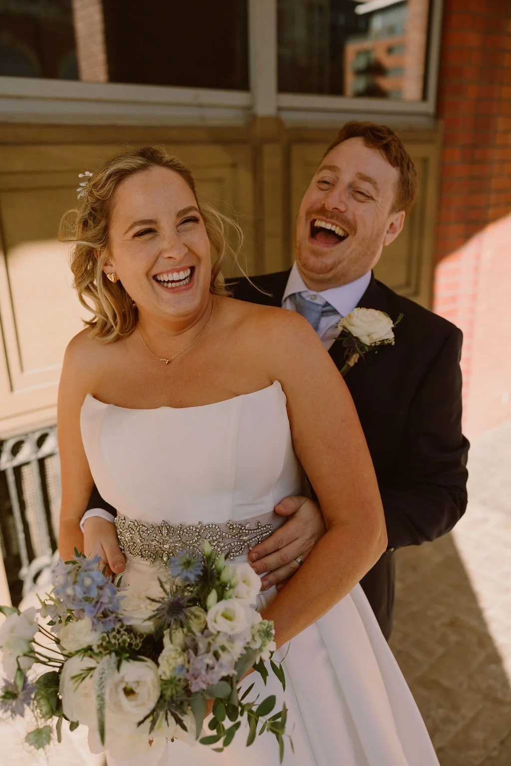 A bride in a white wedding dress holding a bouquet of white and purple flowers, and a groom in a black suit with a white boutonniere, both laughing together.