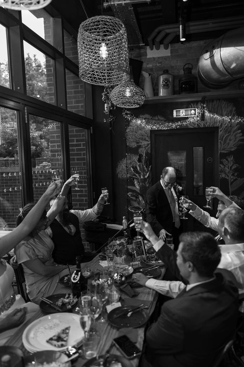 Guests raising glasses in a toast at a dinner party indoors with brick walls, large windows, and decorative lighting.