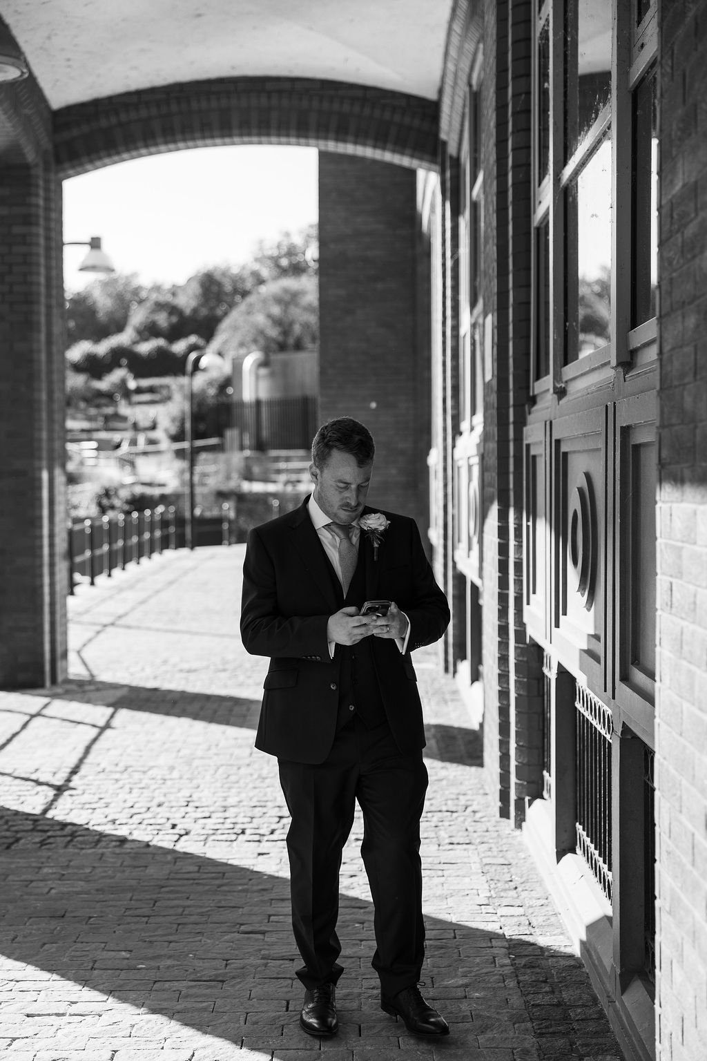A man dressed in a suit with a flower on his lapel standing outdoors under an archway, looking at his phone.