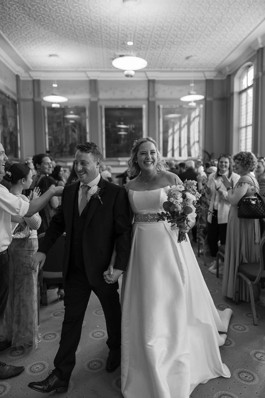 A bride and groom holding hands and smiling at their wedding reception, surrounded by guests clapping and celebrating.