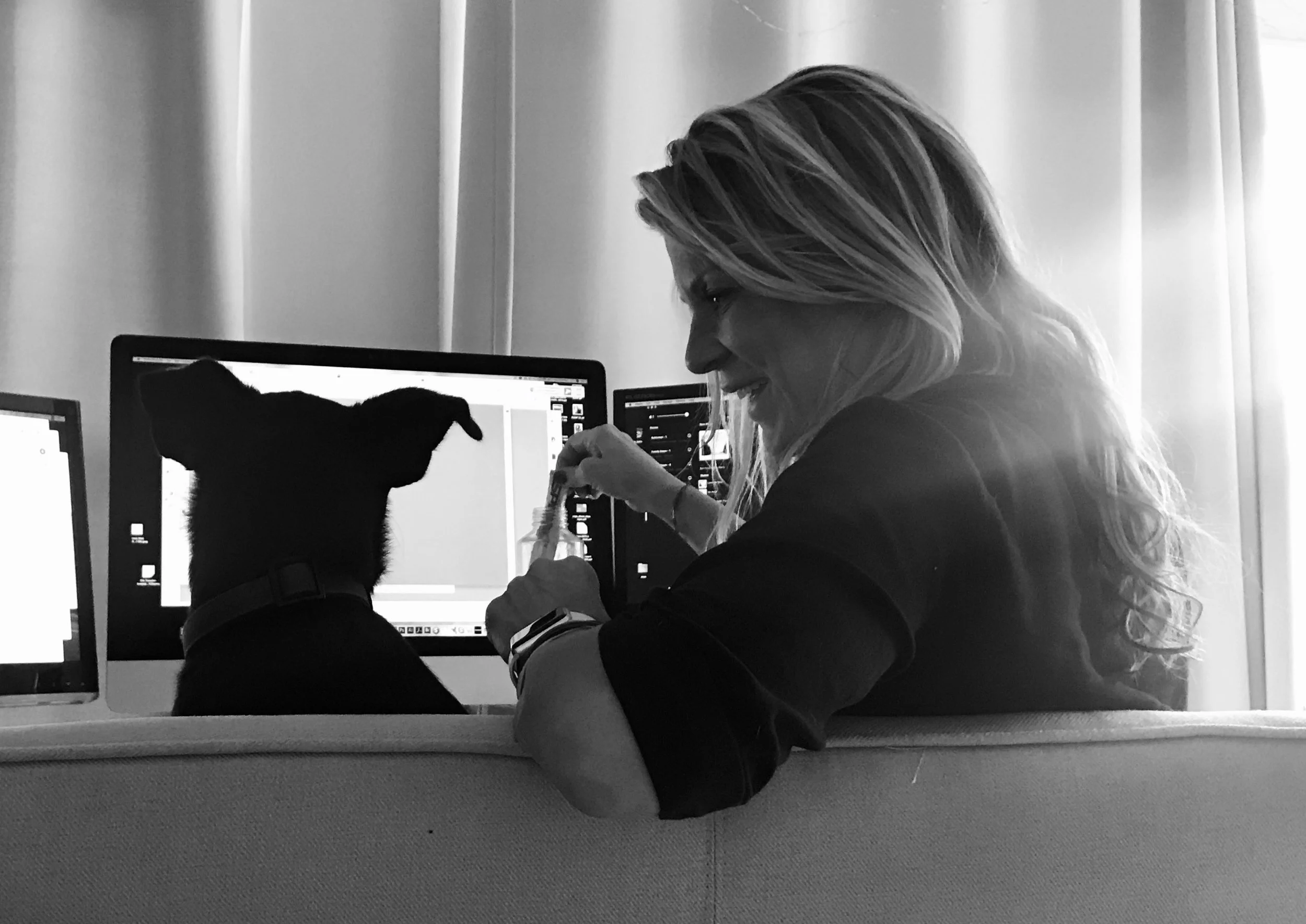 A woman and a dog sitting at a desk with computers, the woman is smiling and appears to be injecting something into the dog.