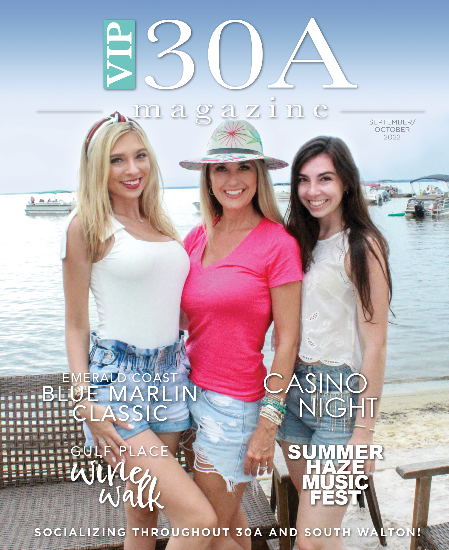 Three women smiling on a beach near the water, with boats in the background. They are dressed in casual summer clothing, with one wearing a colorful hat.