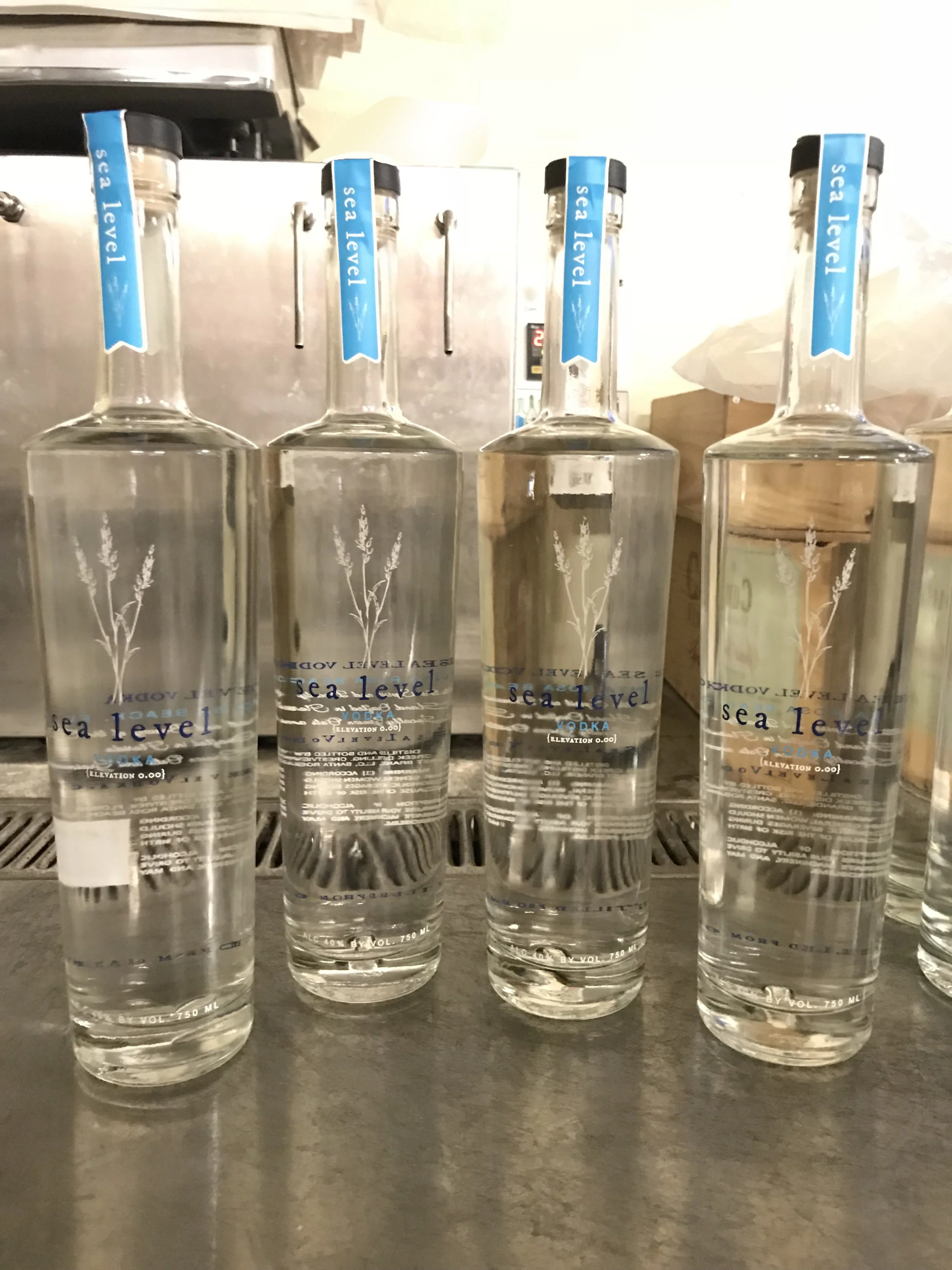 Four bottles of Sea Level vodka on a counter, with dispensers and a kitchen or bar area in the background.
