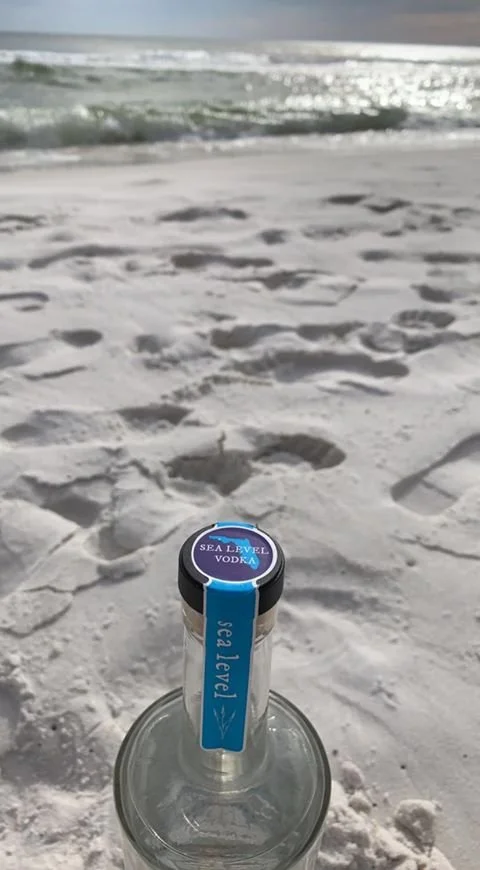 A bottle with a blue label reading 'Sea Level Vodka' on a sandy beach with footprints and ocean waves in the background.