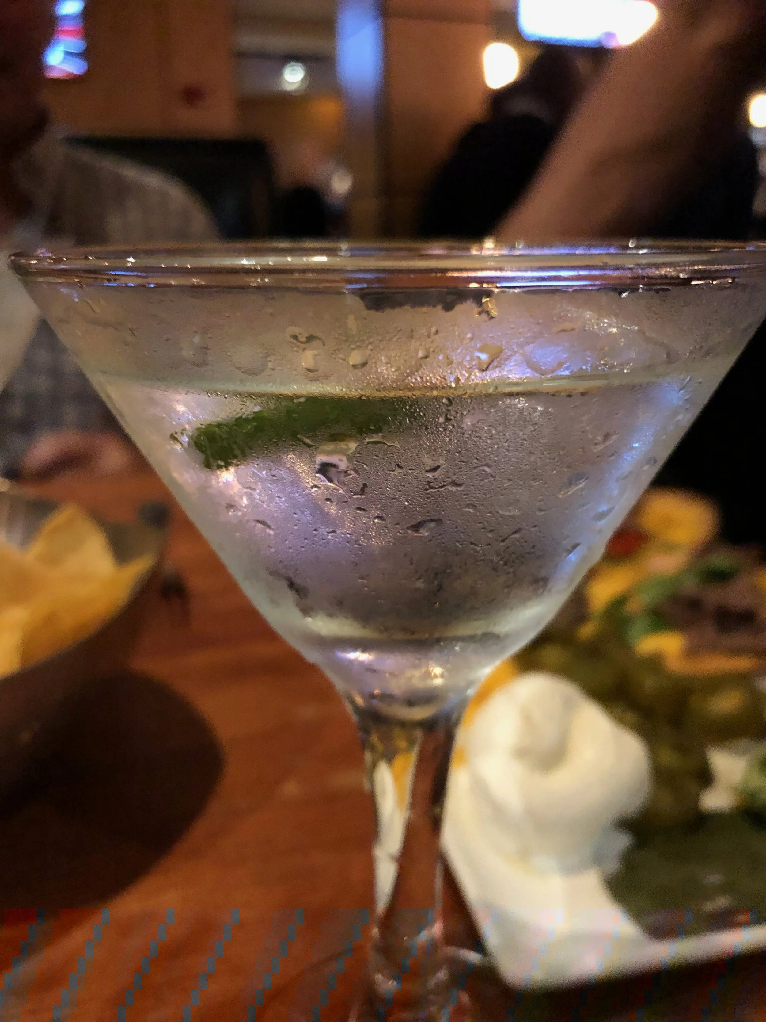 Close-up of a martini glass with a clear cocktail, ice, and lime wedge, condensation on the outside, in a restaurant or bar setting.