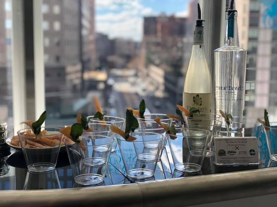 A collection of empty cocktail glasses with garnishes and bottles on a bar counter, overlooking a cityscape through large windows.