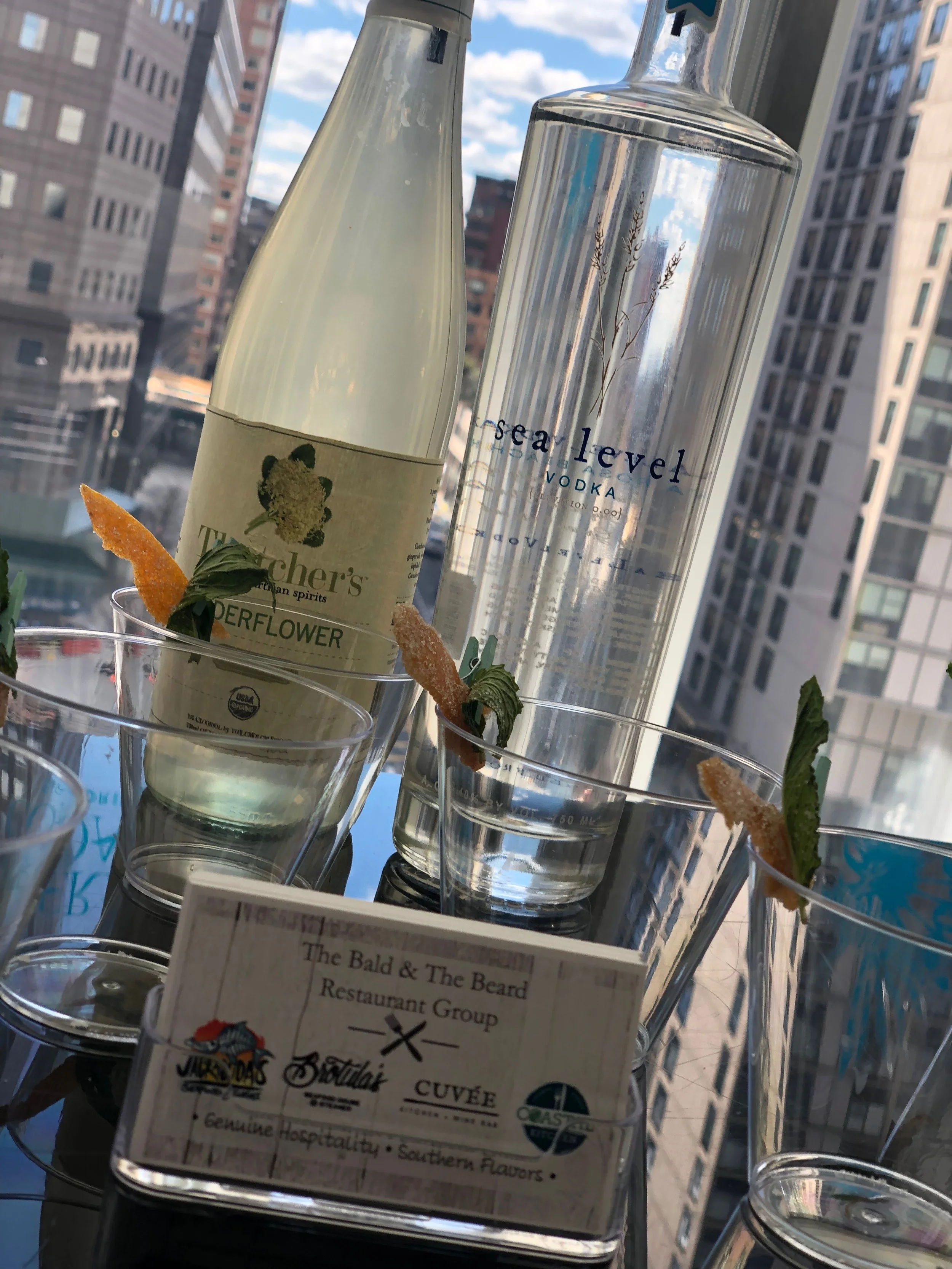 A tall bottle of Tito's Handmade Vodka, a bottle of Sea Level Vodka, and glasses with garnishes on a table. The background shows tall city buildings and a blue sky with clouds.