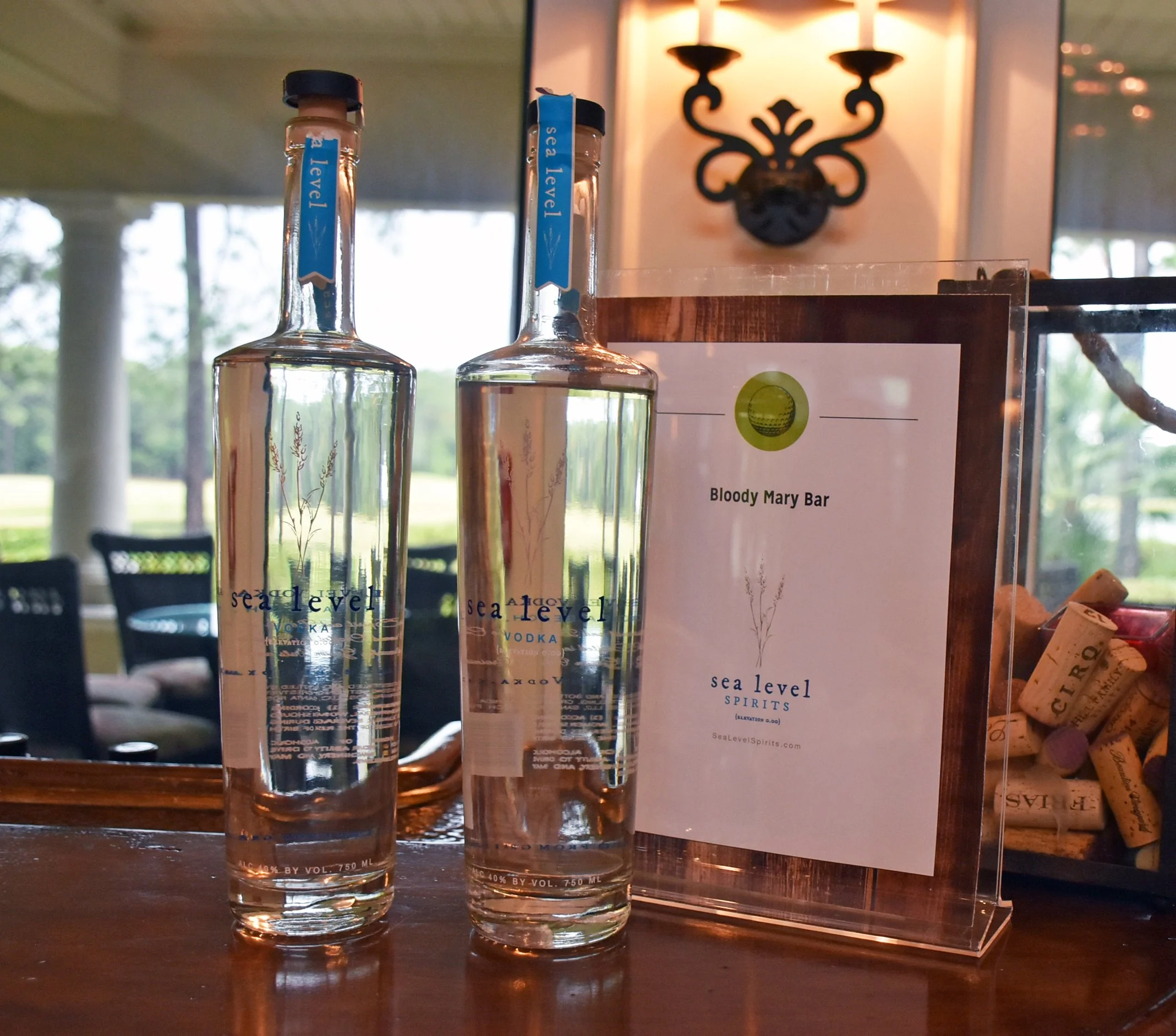 Two bottles of Sea Level vodka and a sign for Bloody Mary Bar at Sea Level Spirits, set on a wooden bar counter with a window and outdoor scenery in the background.