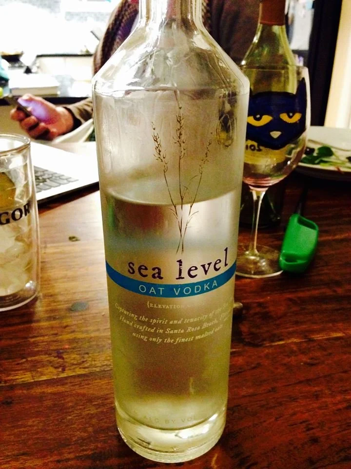 A tall, clear bottle of Sea Level Oat Vodka with a plant illustration on the label, partially filled with liquid, placed on a wooden table with other drinks and a person in the background.