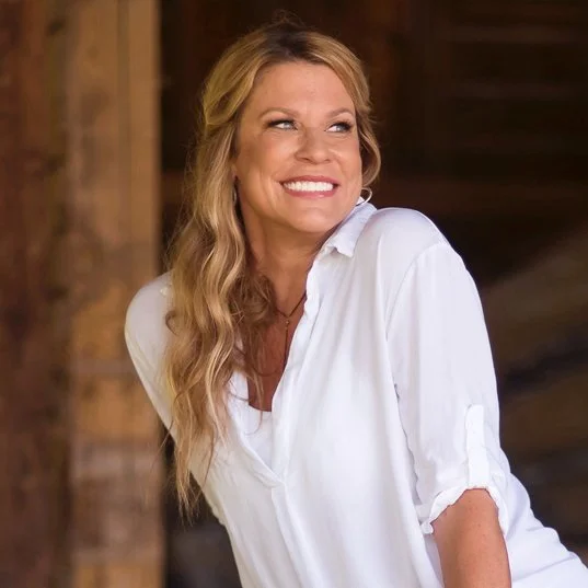 A woman with long wavy blonde hair smiling and looking to her right, wearing a white shirt, standing outdoors near a wooden structure.