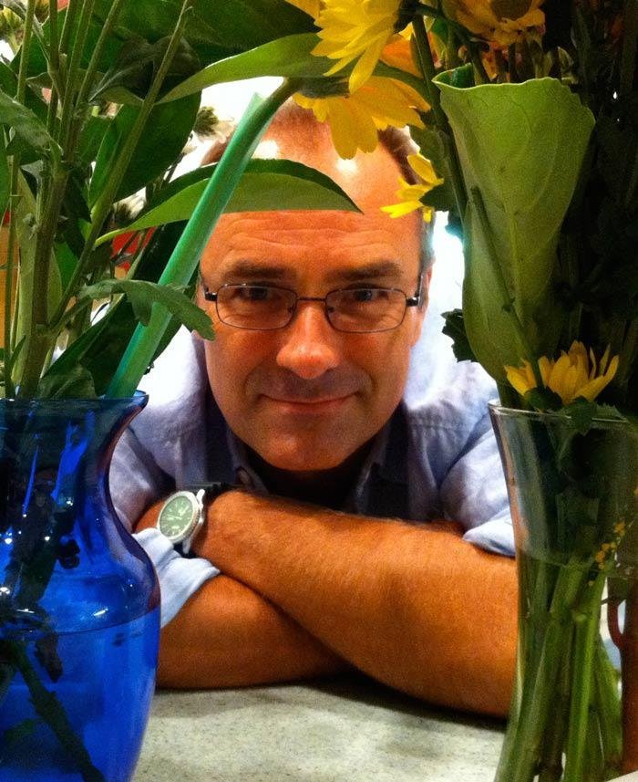 A man with glasses lying on a table, surrounded by colorful flower vases, looking at the camera.
