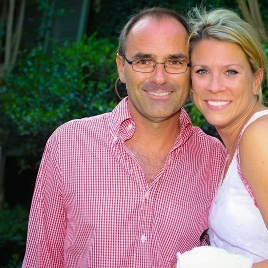 A smiling man and woman standing close together outdoors, with greenery in the background.