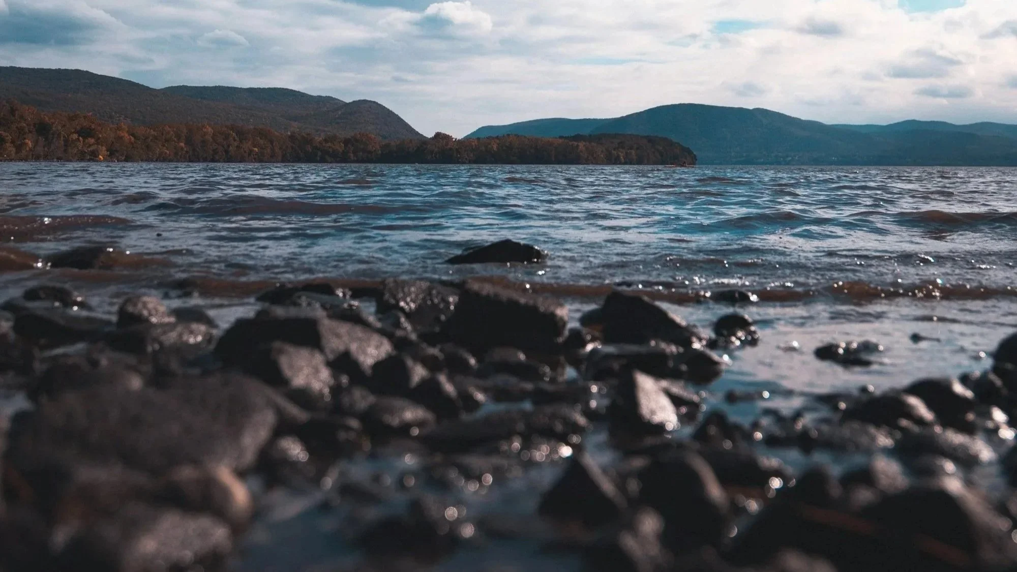 Close-up view of rocky shore at a lake or river, with water in the foreground and hills in the background under a cloudy sky.