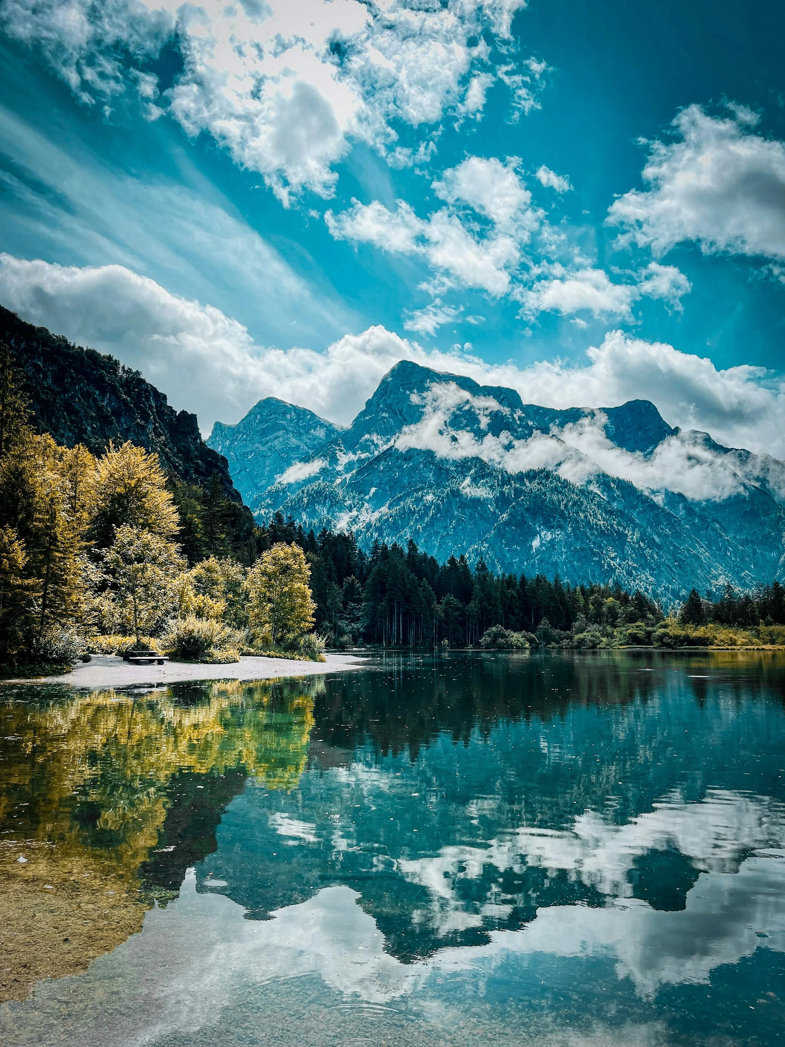 The image showcases a serene mountain landscape reflecting in a calm lake. It features a backdrop of towering, snow-capped mountains under a partly cloudy sky. The foreground includes a shoreline with trees and green foliage.