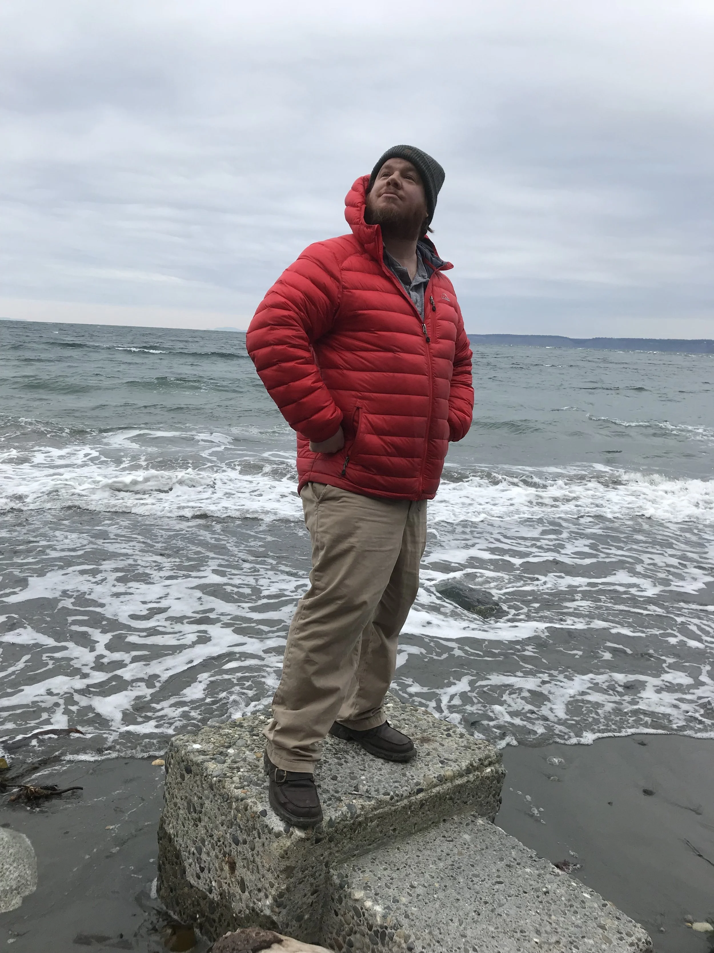 A man standing on a large rock on the shore, facing the water with his hands in his jacket pockets. He is wearing a red puffy jacket, tan pants, a beanie, and hiking shoes, with the ocean and cloudy sky in the background.