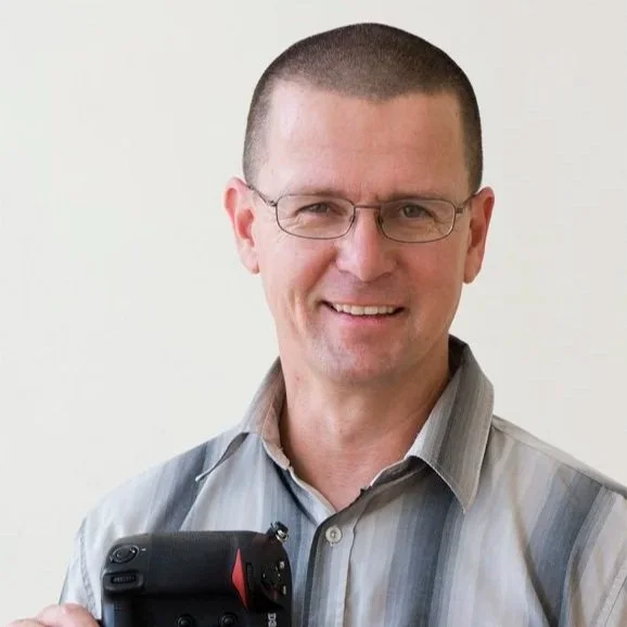 Smiling man with glasses and short hair holding a camera, wearing a striped button-up shirt, against a plain white background.