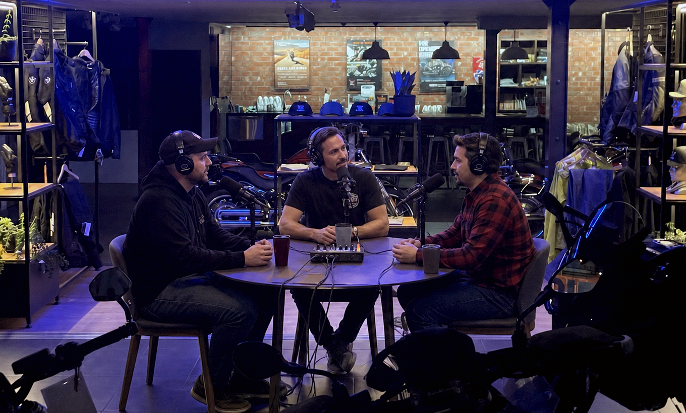 Three men sit at a round table in a podcast studio, speaking into microphones, all wearing headphones. The studio has a brick wall background with clothing racks and motorcycle helmets on shelves. The scene is dimly lit with ceiling and background lighting.