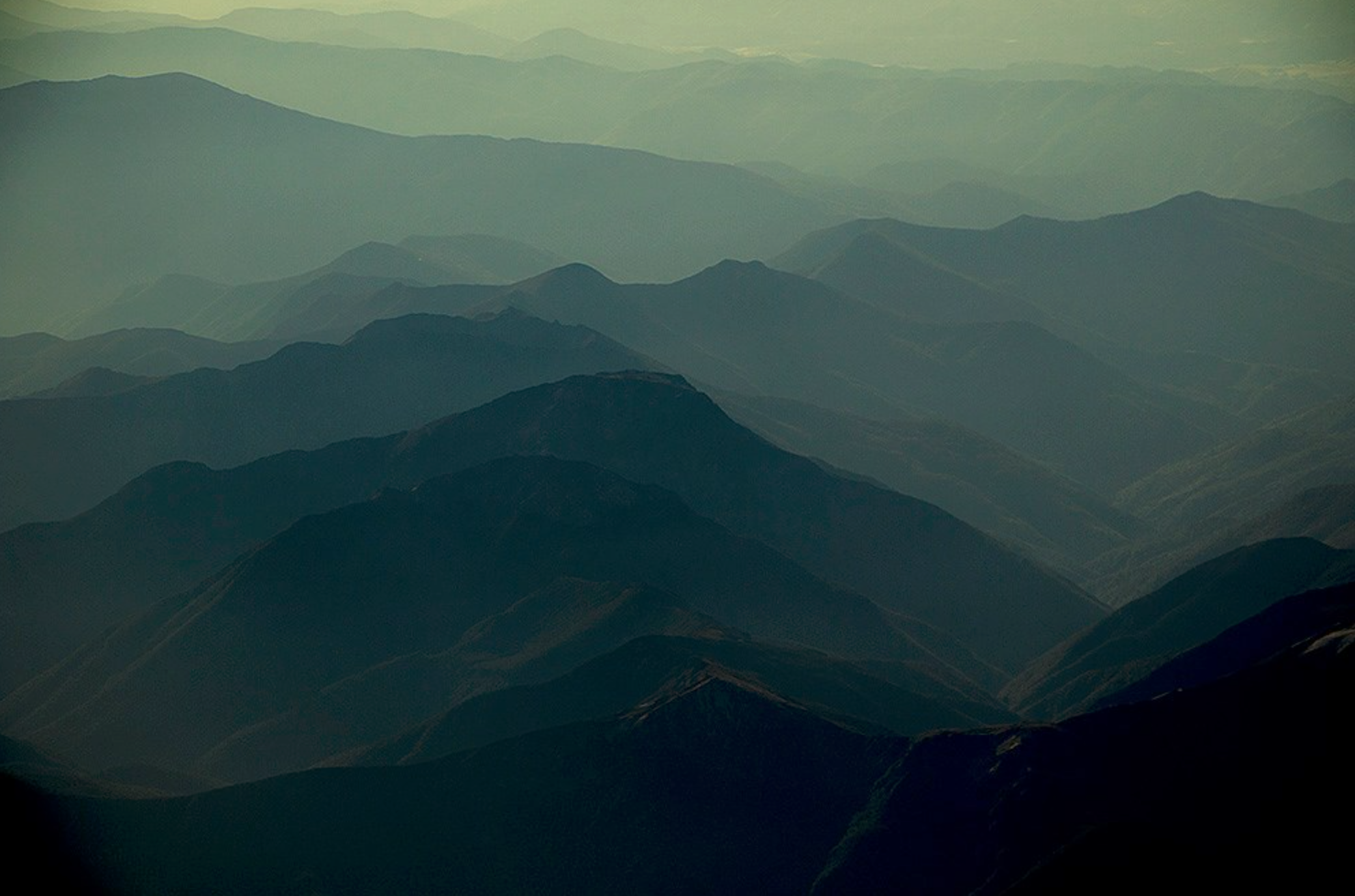 Aerial view of layered mountains with a greenish haze, showing multiple ridges and peaks receding into the distance.