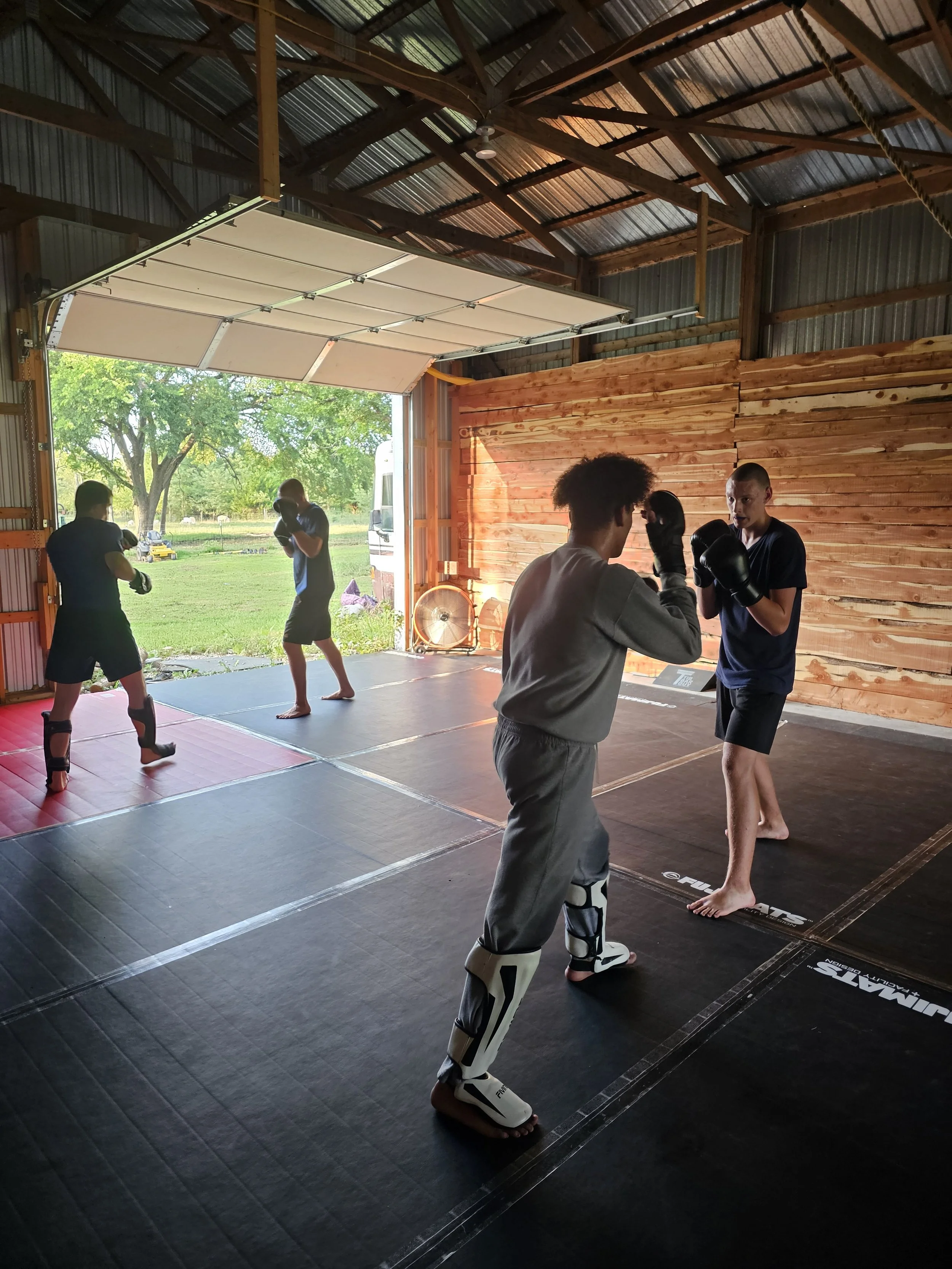 Jiu Jitsu, Grapplina, MMA, Muay Thai, Group Classes, BJJ
Bourbon County Fight Factory, Fort Scott, KS Boxers practicing fighting in a gym with wooden wall and open garage door view of greenery outside