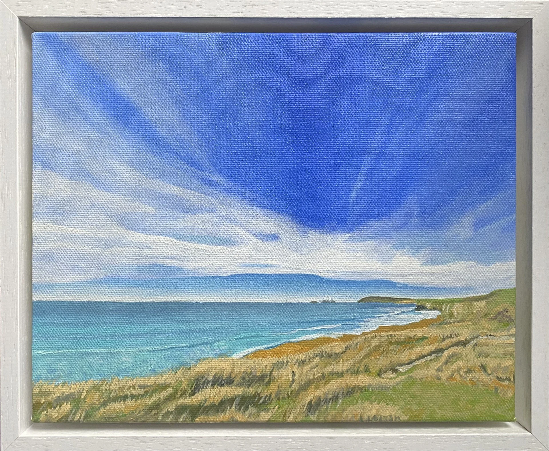 A framed painting of a Cornish coastal scene with Godrevy Lighthouse of Virginia Woolf's novel. Blue sky, white clouds, ocean waves, and sandy beach with grass and rocks in the foreground.