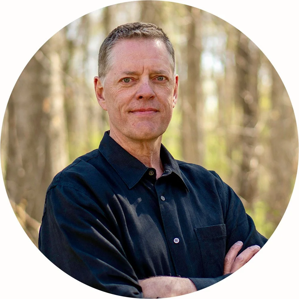 A man in a dark button-up shirt standing outdoors with trees in the background, arms crossed, looking at the camera.