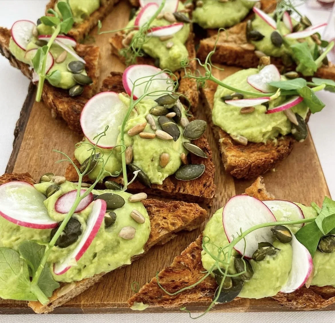 Toasted bread slices topped with mashed avocado, radish slices, sunflower seeds, and microgreens on a wooden platter.