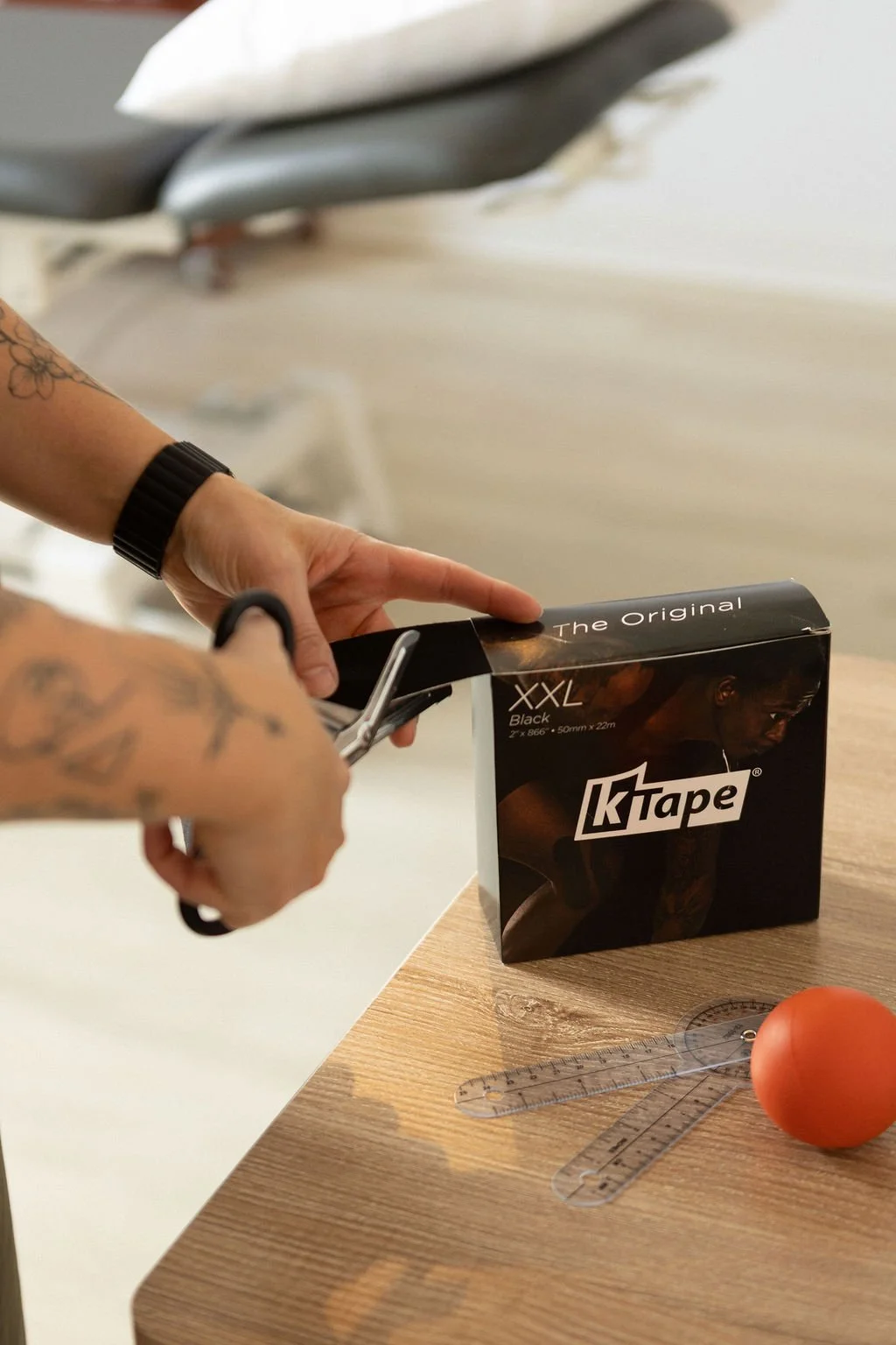 A person with tattoos on their arms is using scissors to cut a black box of XXL black KTAPE tape. The scene is set on a wooden table with a plastic goniometer, a ruler, and an orange ball nearby.