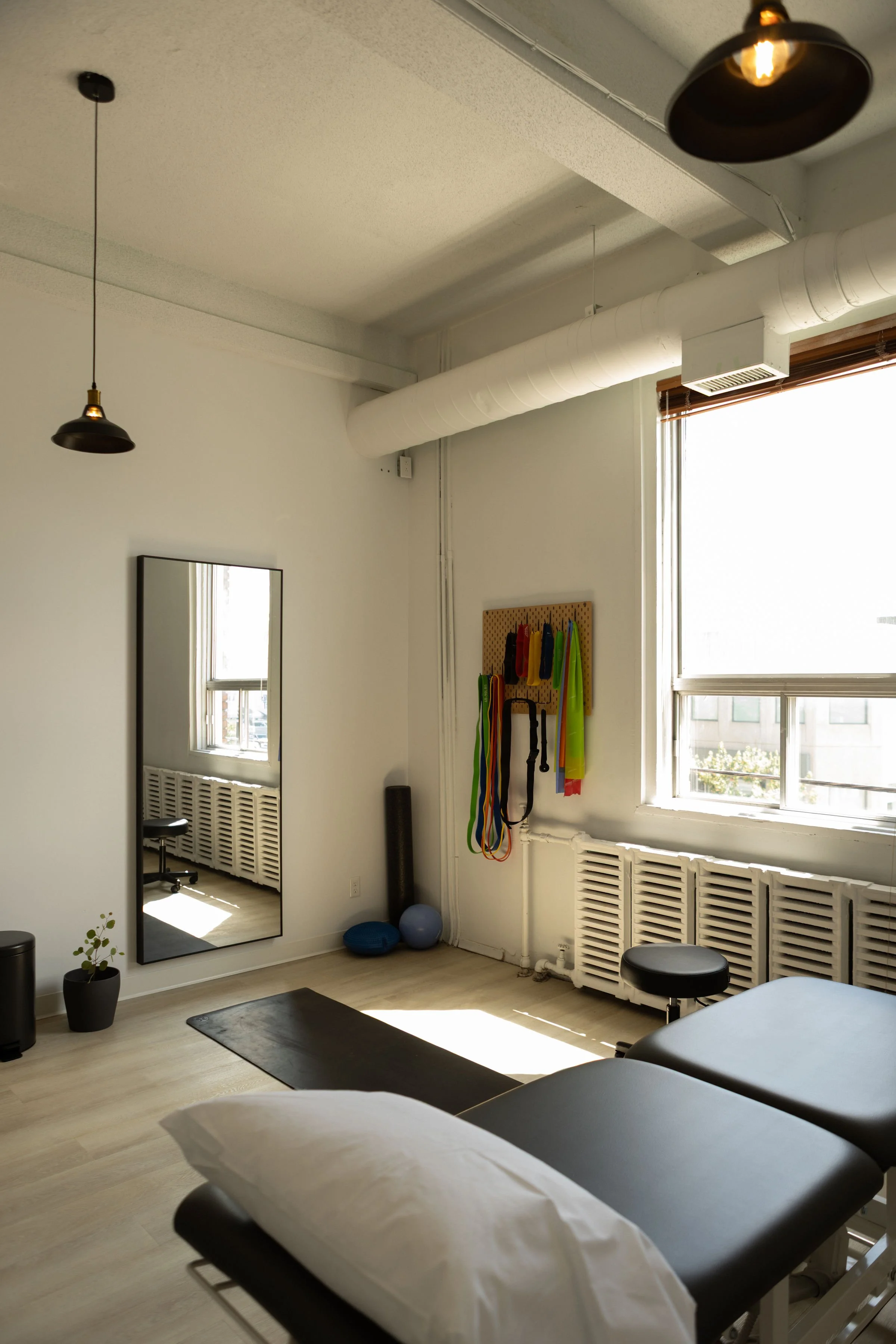 A room with therapy table, two pillows with white sheets, a mirror, exercise bands hanging on a pegboard, exercise balls, a foam roller, a plant in a pot, large windows with blinds and curtains, and hanging pendant lights.