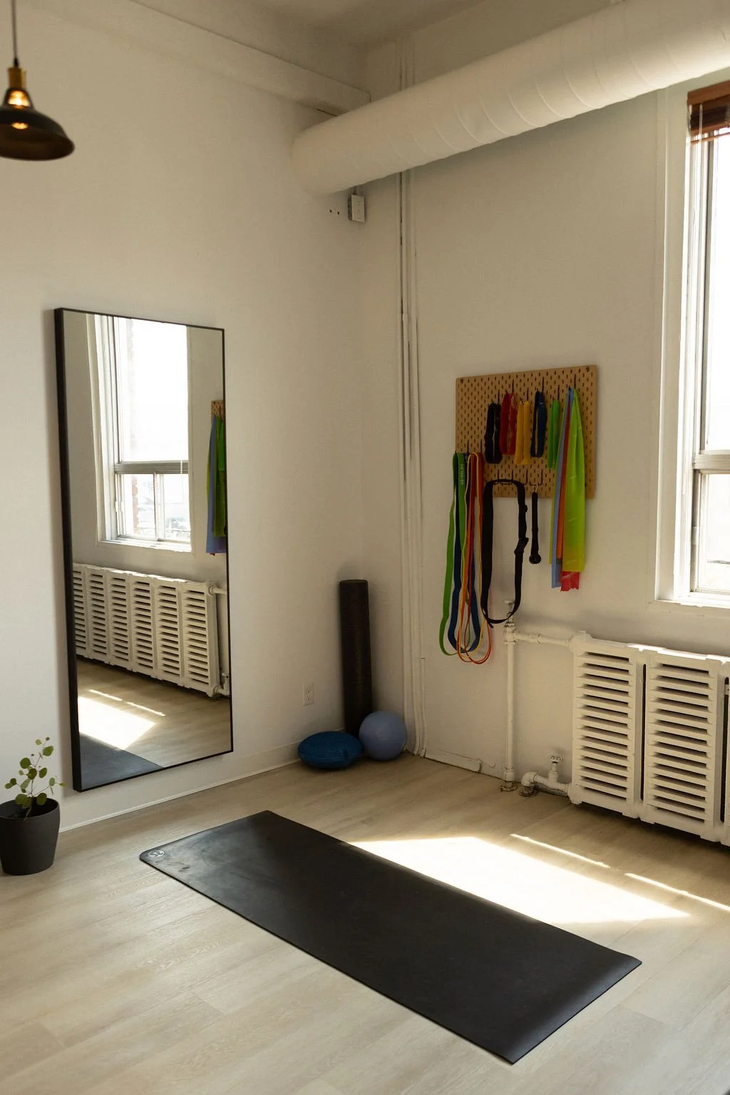 Yoga or workout studio with black mat, mirror, radiator, window, and athletic equipment like resistance bands, balls, and foam roller.