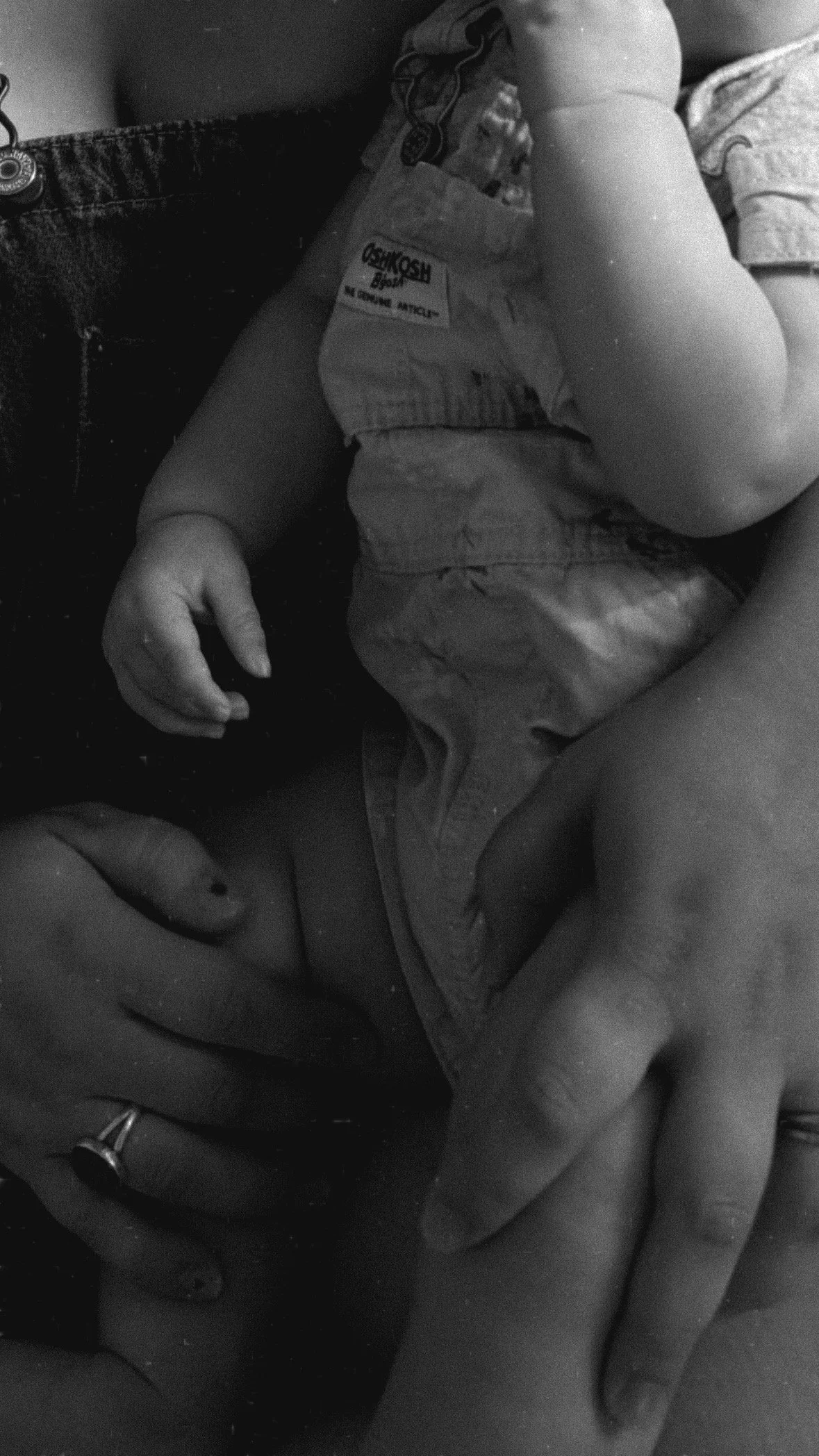 A small child in a diaper and shirt, sitting with one leg crossed over the other, looking down, with hands resting on their legs. Face is not visible.