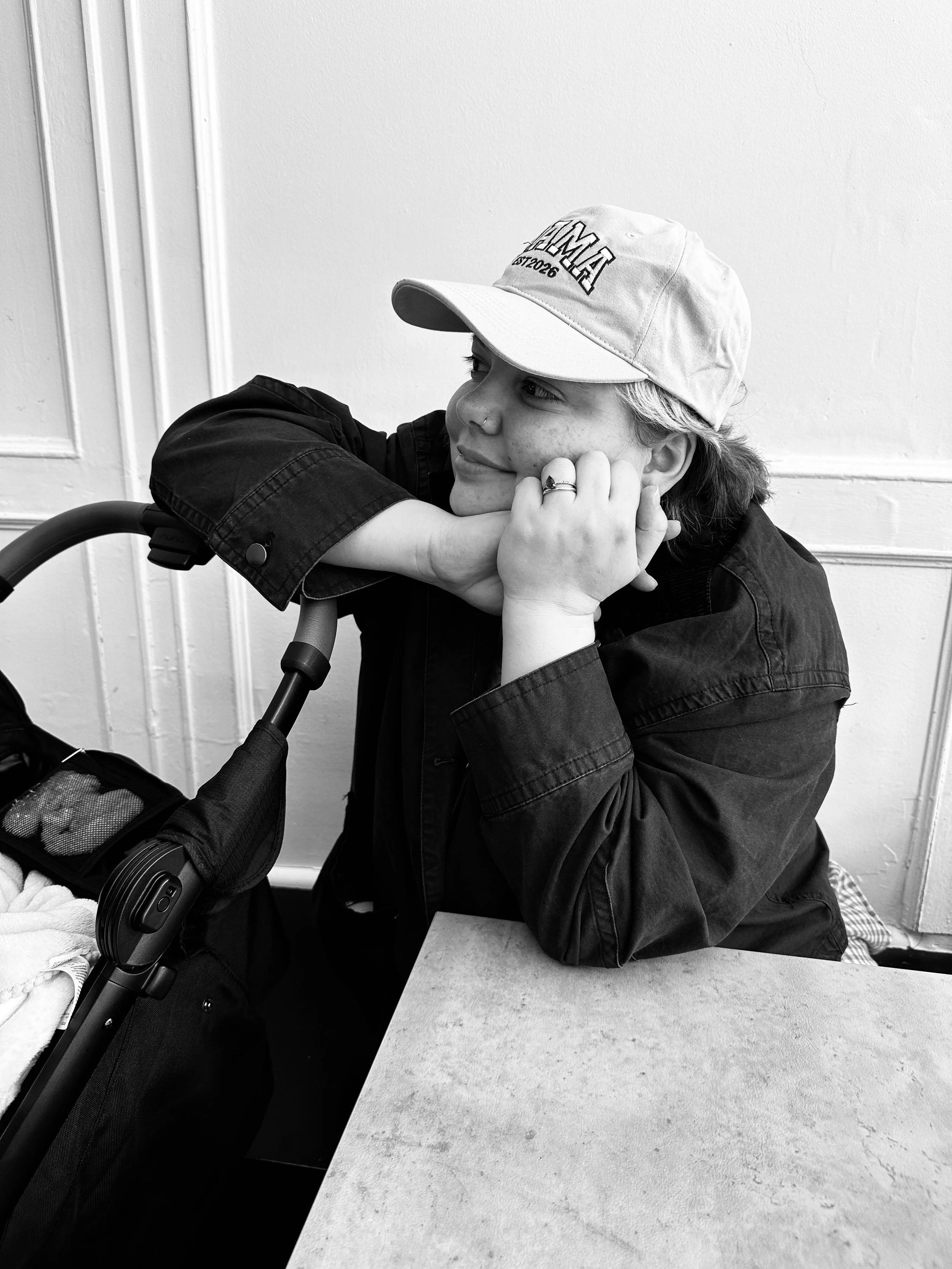 Black and white photo of a white woman leaning on a baby stroller, wearing a cap that says "MAMA, Est. 2026" and a jacket, resting her chin on her folded arms on a table, smiling and looking to her right.