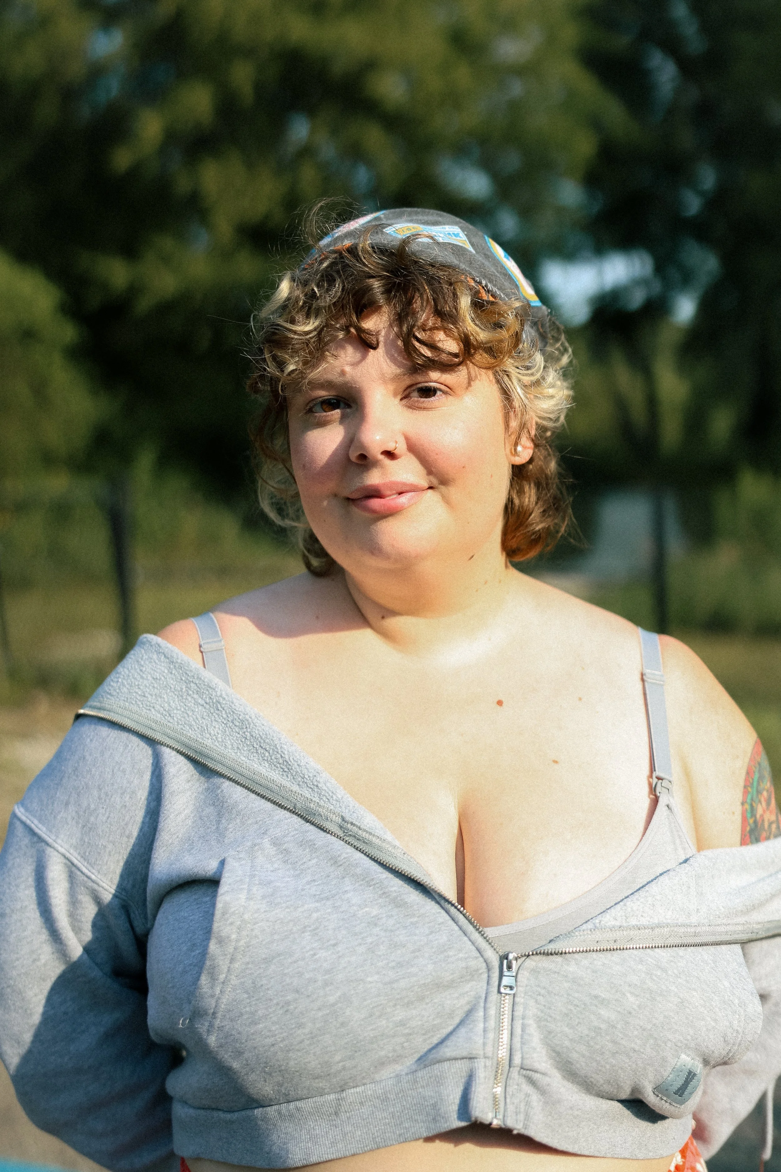 A young white woman with short curly hair and a nose piercing smiling outdoors. She is wearing a gray zip-up hoodie partially off her shoulders and a black patterned bandana. The background is blurred trees and sky.
