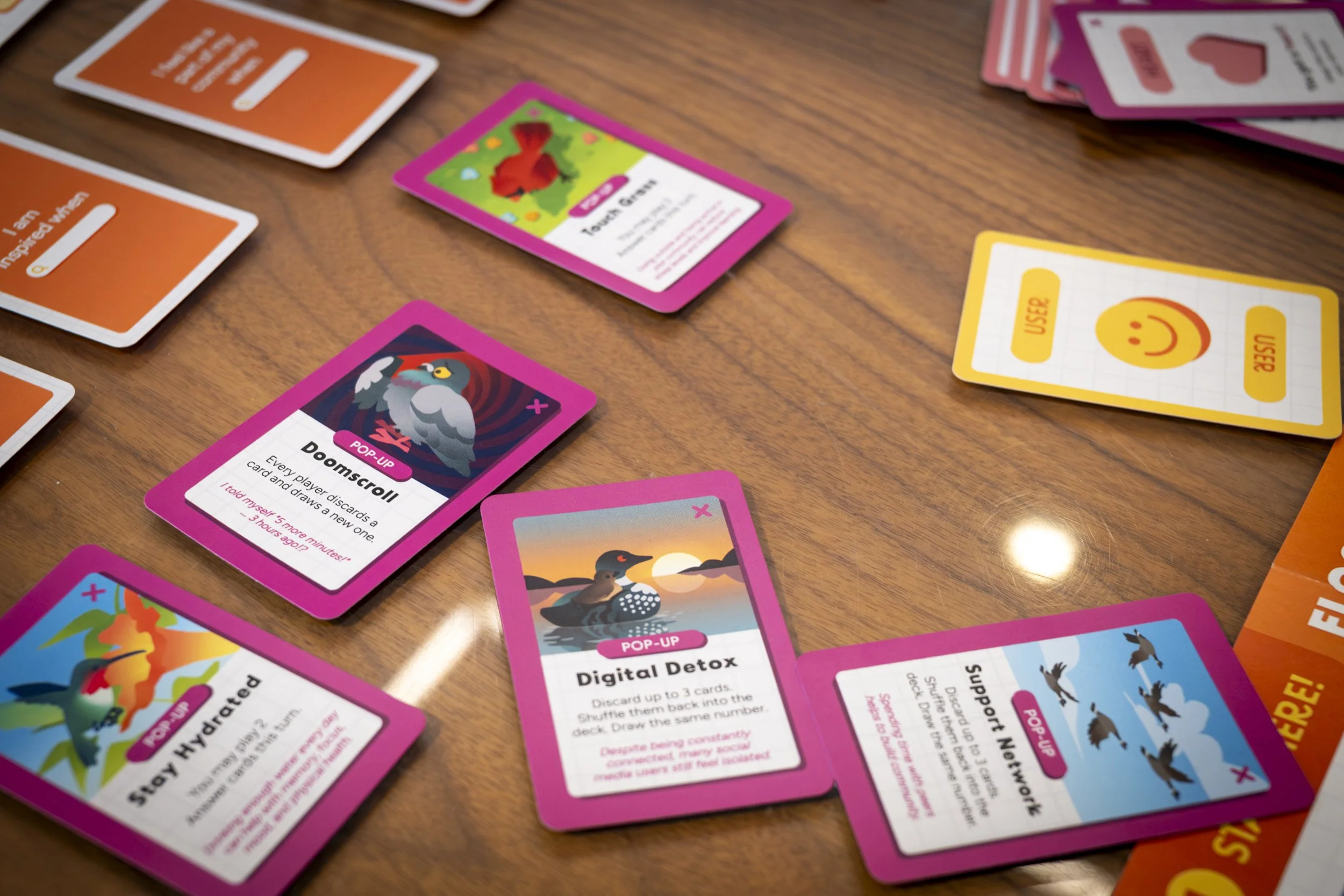 Colorful game cards scattered on a wooden table, including cards labeled Doomscroll, Digital Detox, Stay Hydrated, Support Network, and user and use cards with a smiley face.