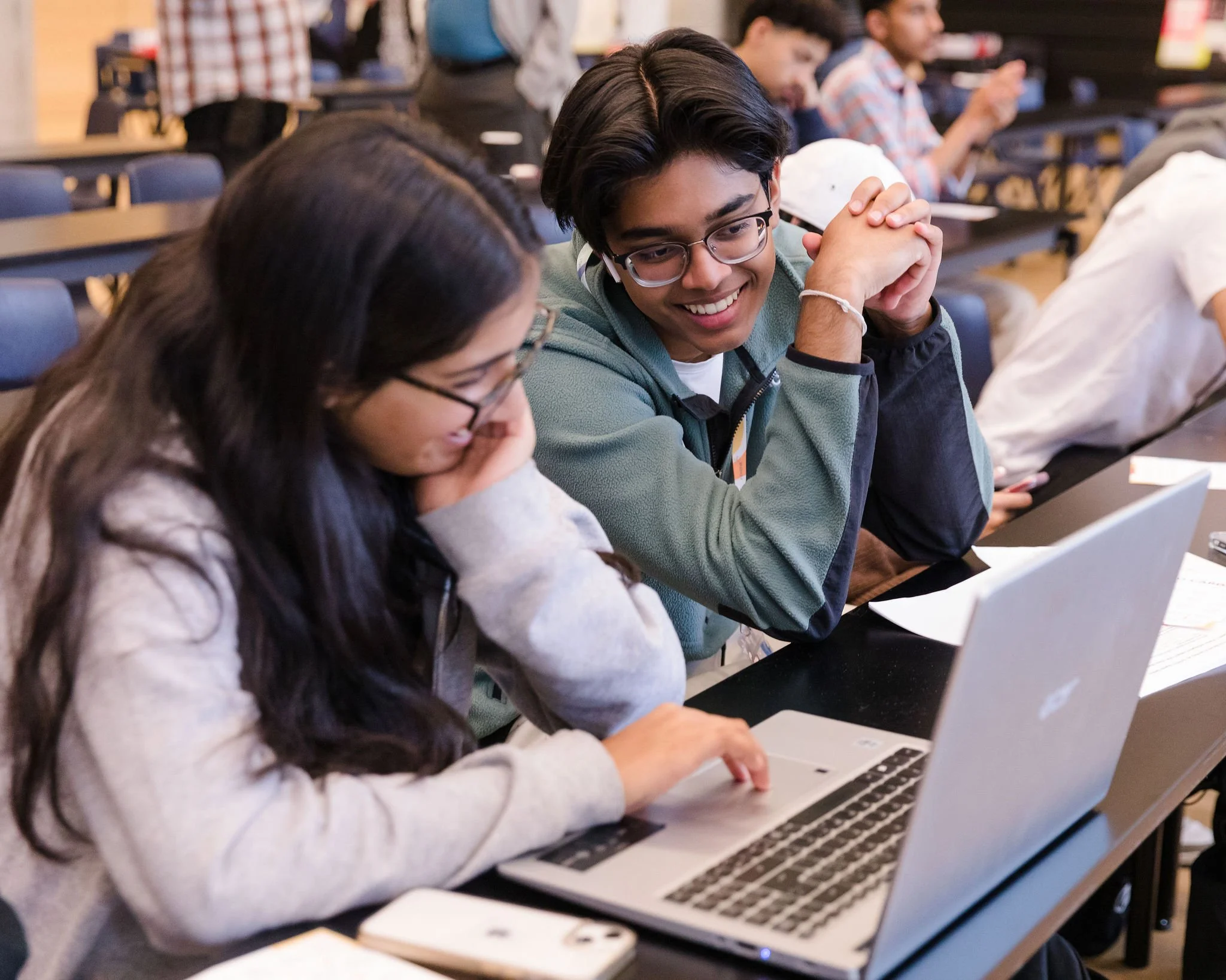 Two students, a girl and a boy, sitting at a desk looking at a laptop together in a classroom or conference setting, with other students in the background.