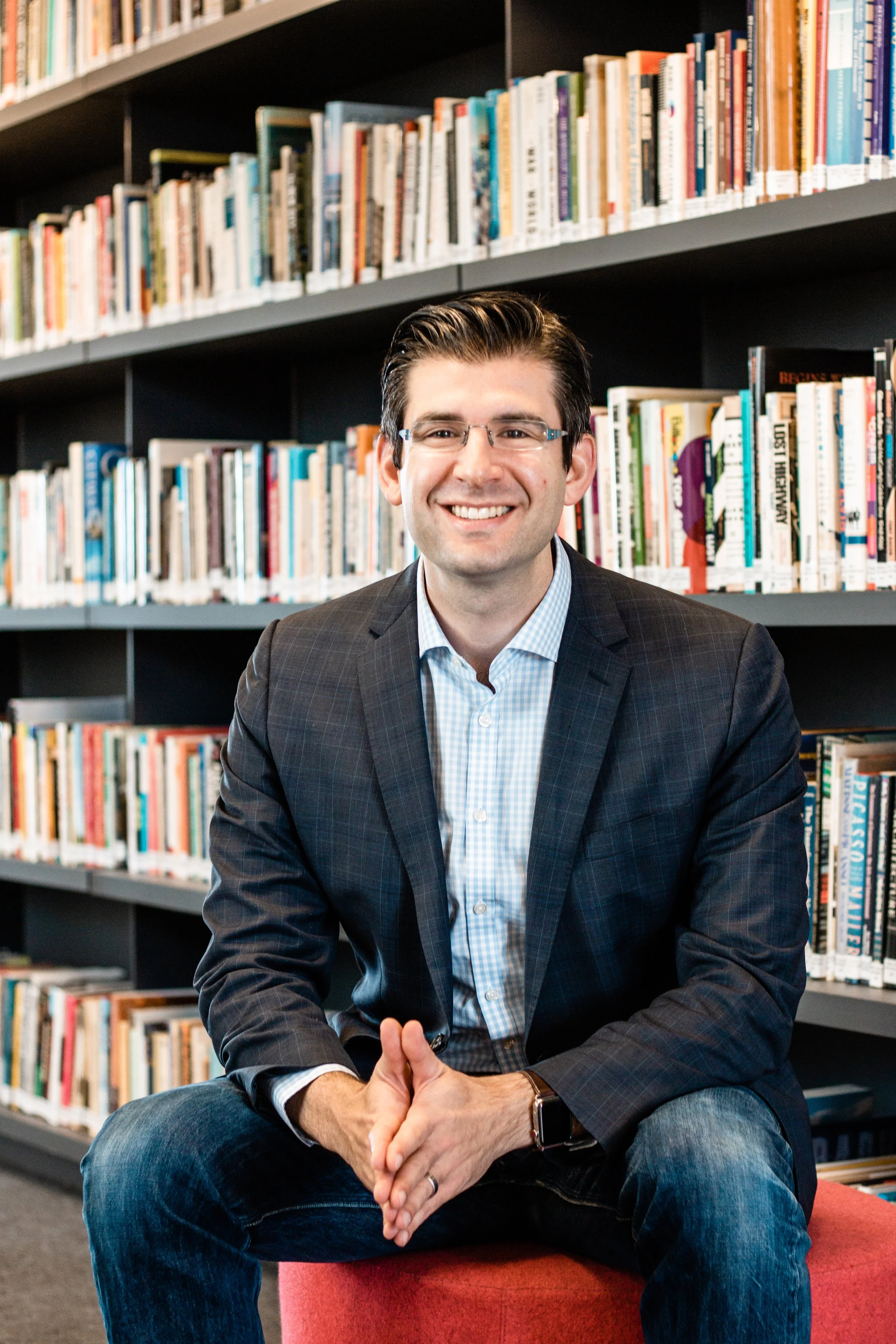 A smiling man with glasses, wearing a blazer and jeans, sitting on a red stool in front of bookshelves in a library.