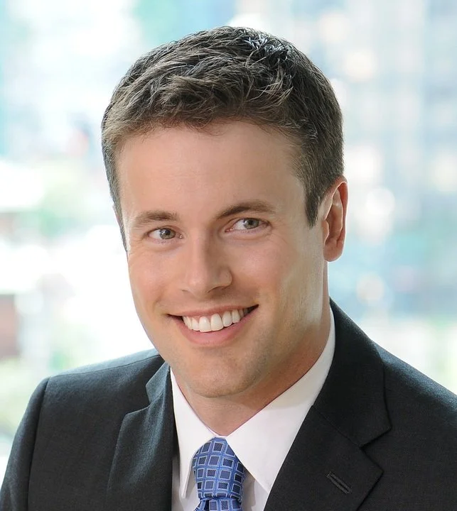 Professional headshot of a young man in a suit and tie, smiling with a cityscape background.